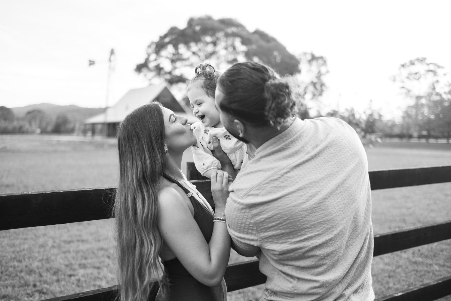 Parents share a tender moment kissing their baby at golden hour on a farm in black and white portrait.