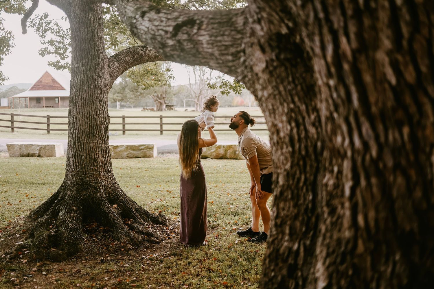 Family of three playing with baby under large trees on a farm with white barn background.