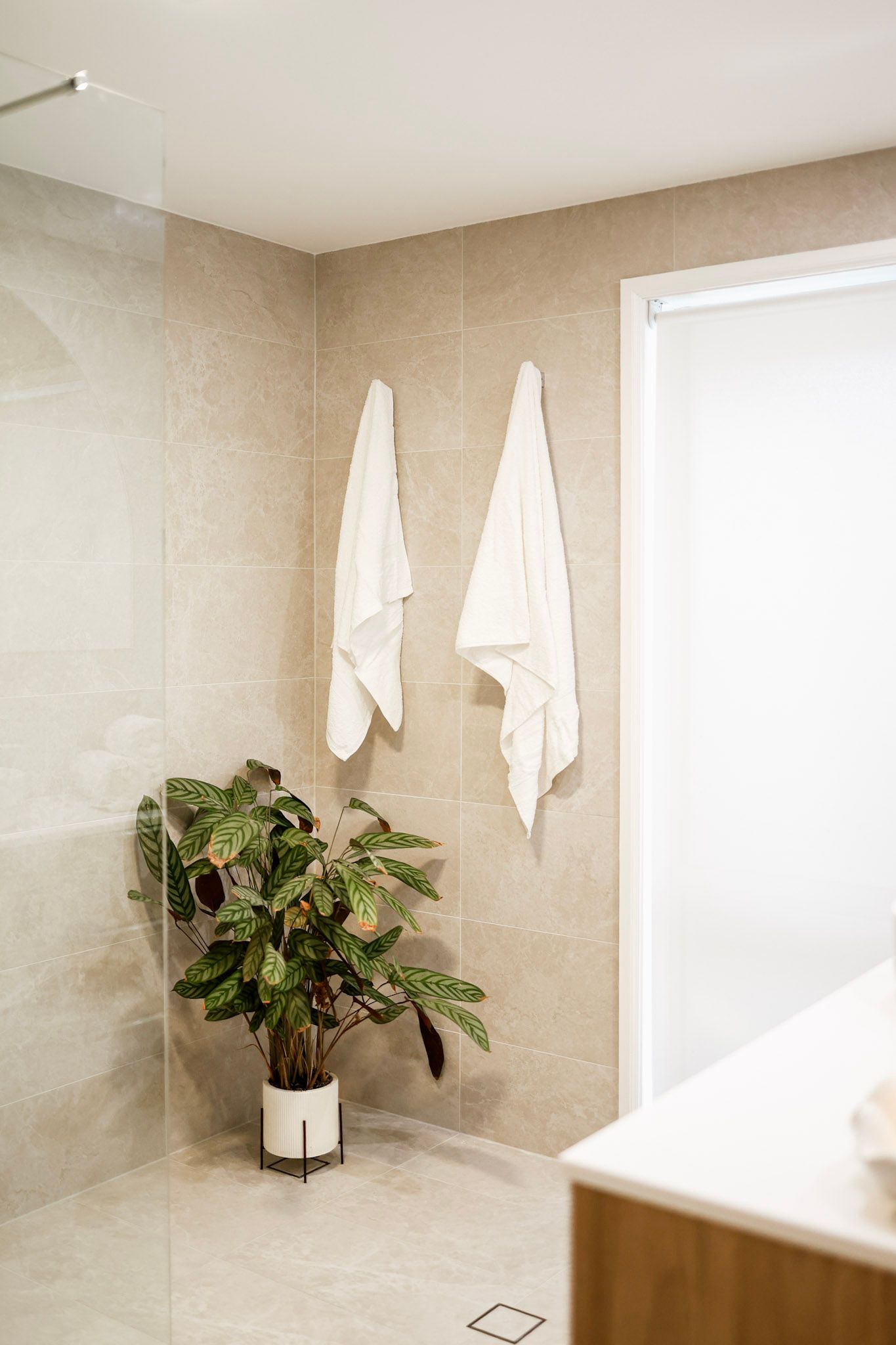 Modern bathroom with beige tiles, two white towels on hooks, and a green potted plant in the corner.