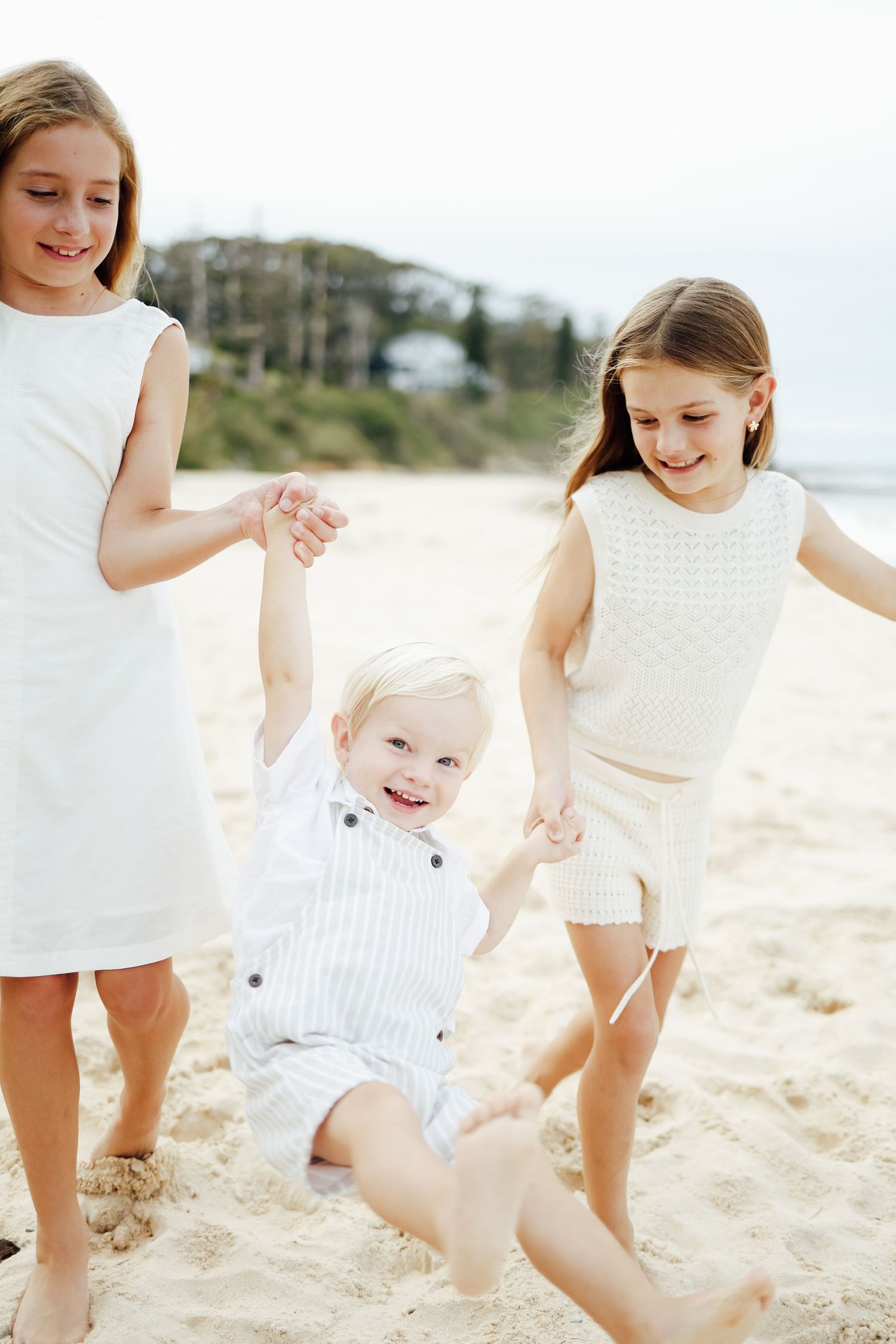 Two girls in white dresses swinging a laughing toddler boy by his hands on a sandy beach.