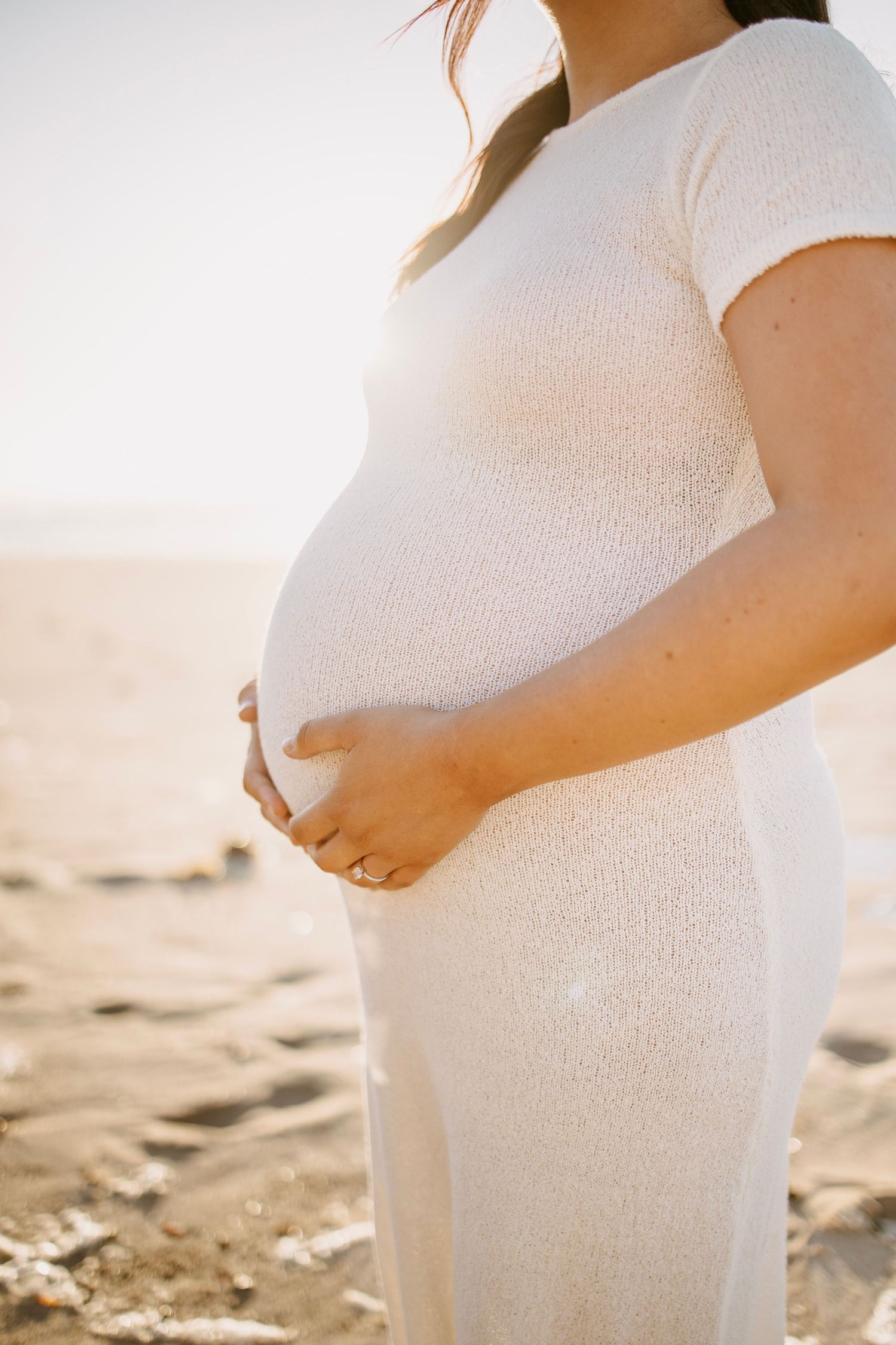 Pregnant woman in white dress cradling baby bump on sunny sandy beach.