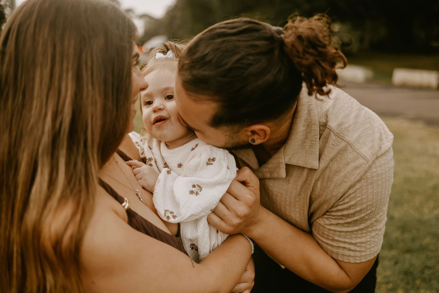 Parents lovingly kissing their smiling baby outdoors in soft natural bokeh light.