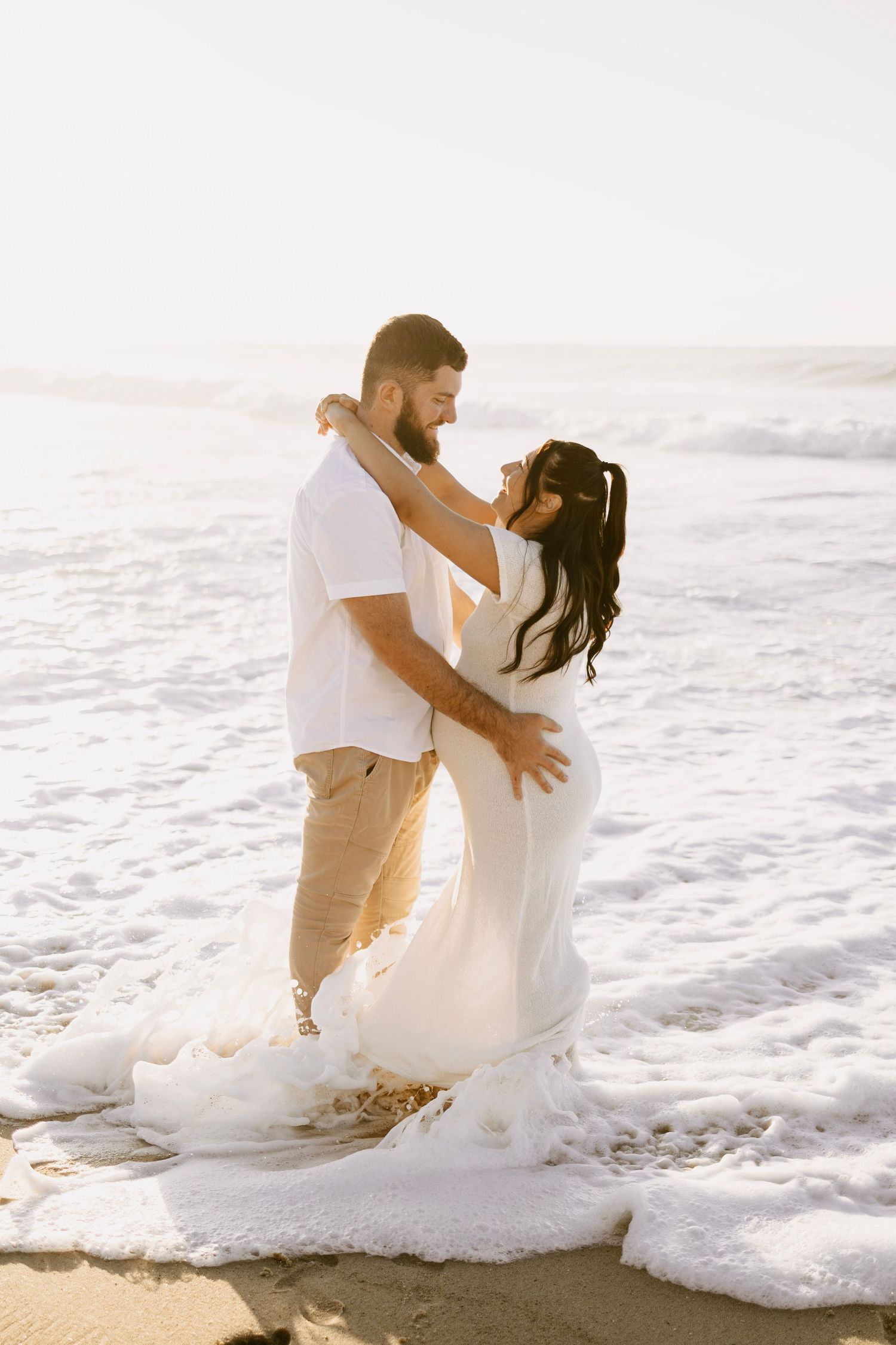 Couple embracing in ocean waves at sunset, woman in flowing white gown, man in white shirt and tan pants.