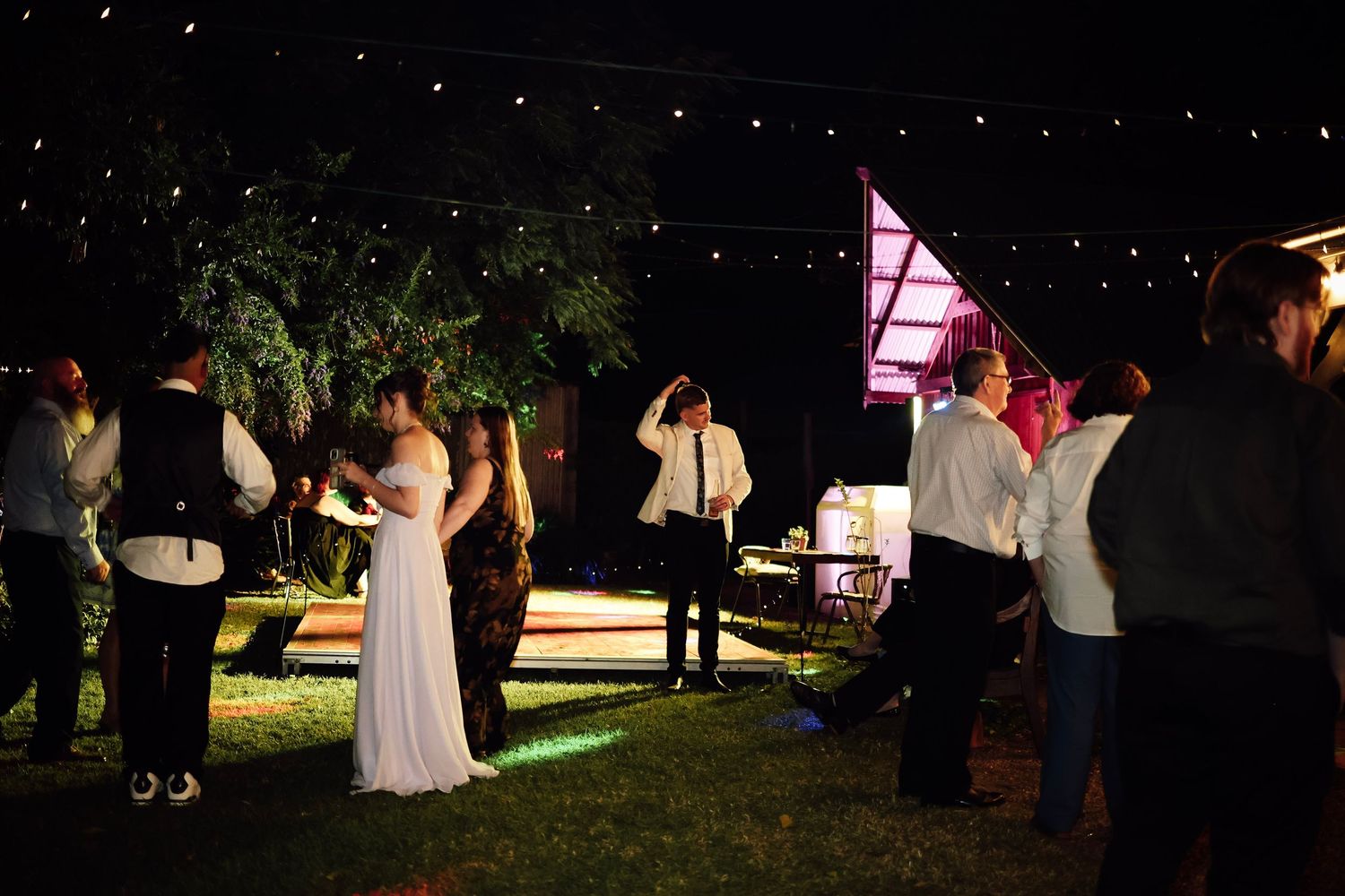 Bride and groom dancing outdoors at a night wedding reception with string lights and guests surrounding them.