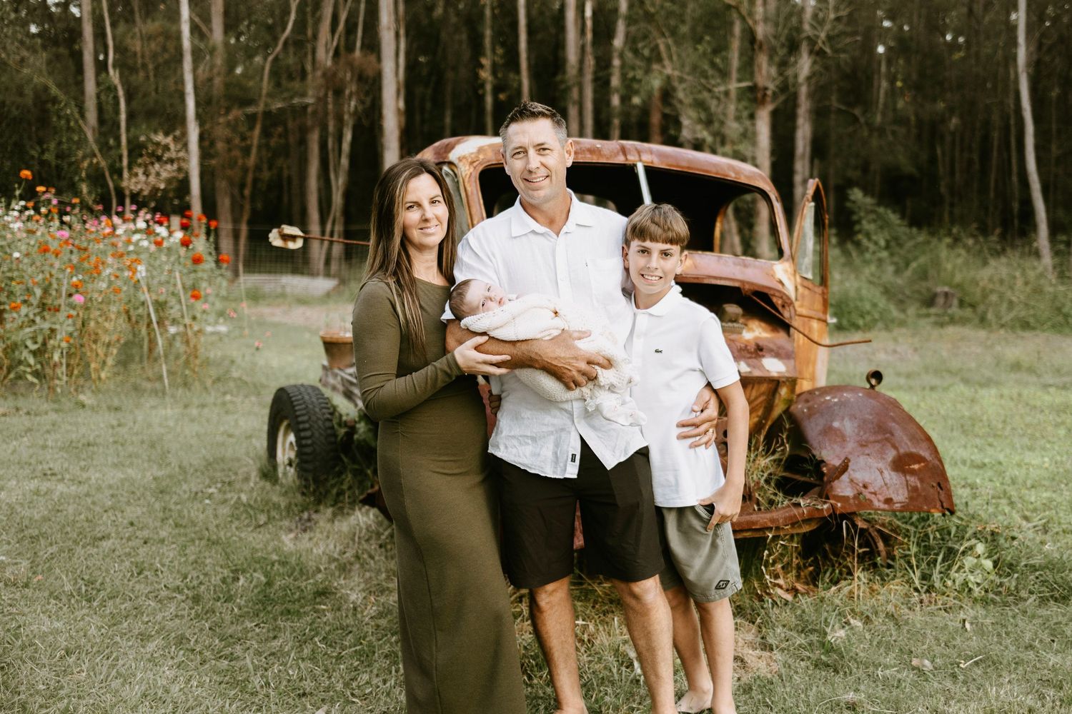 A family of four poses in front of a rustic vintage truck surrounded by wildflowers in a field.