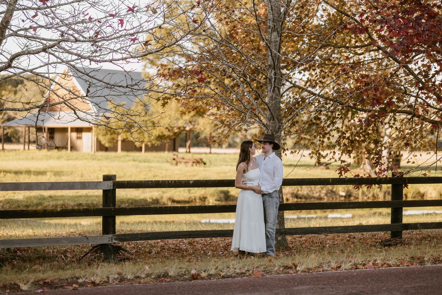 Romantic couple embracing under a colorful autumn tree beside a wooden fence on a farm.