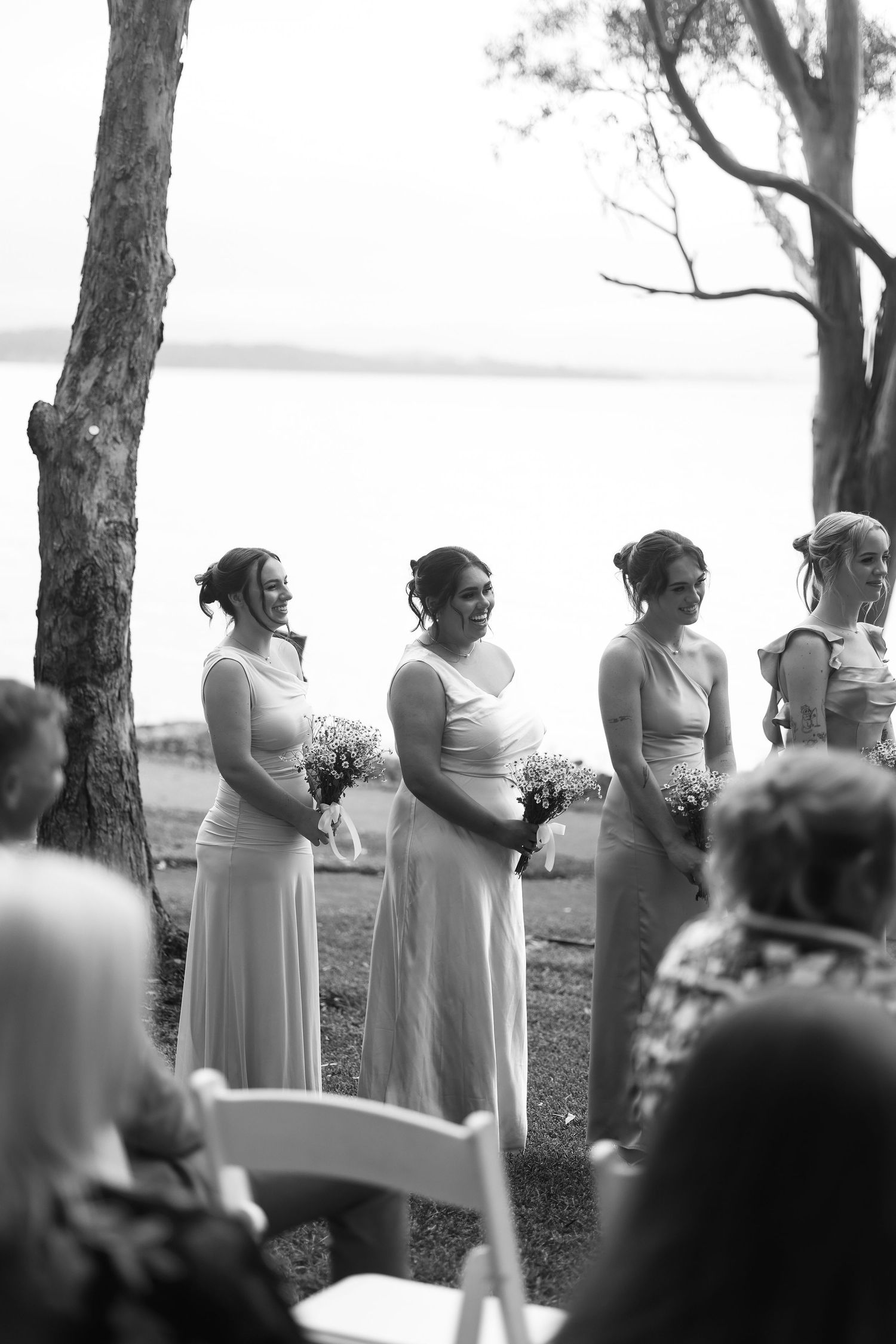 Four bridesmaids in white dresses holding bouquets at an outdoor waterside wedding ceremony.
