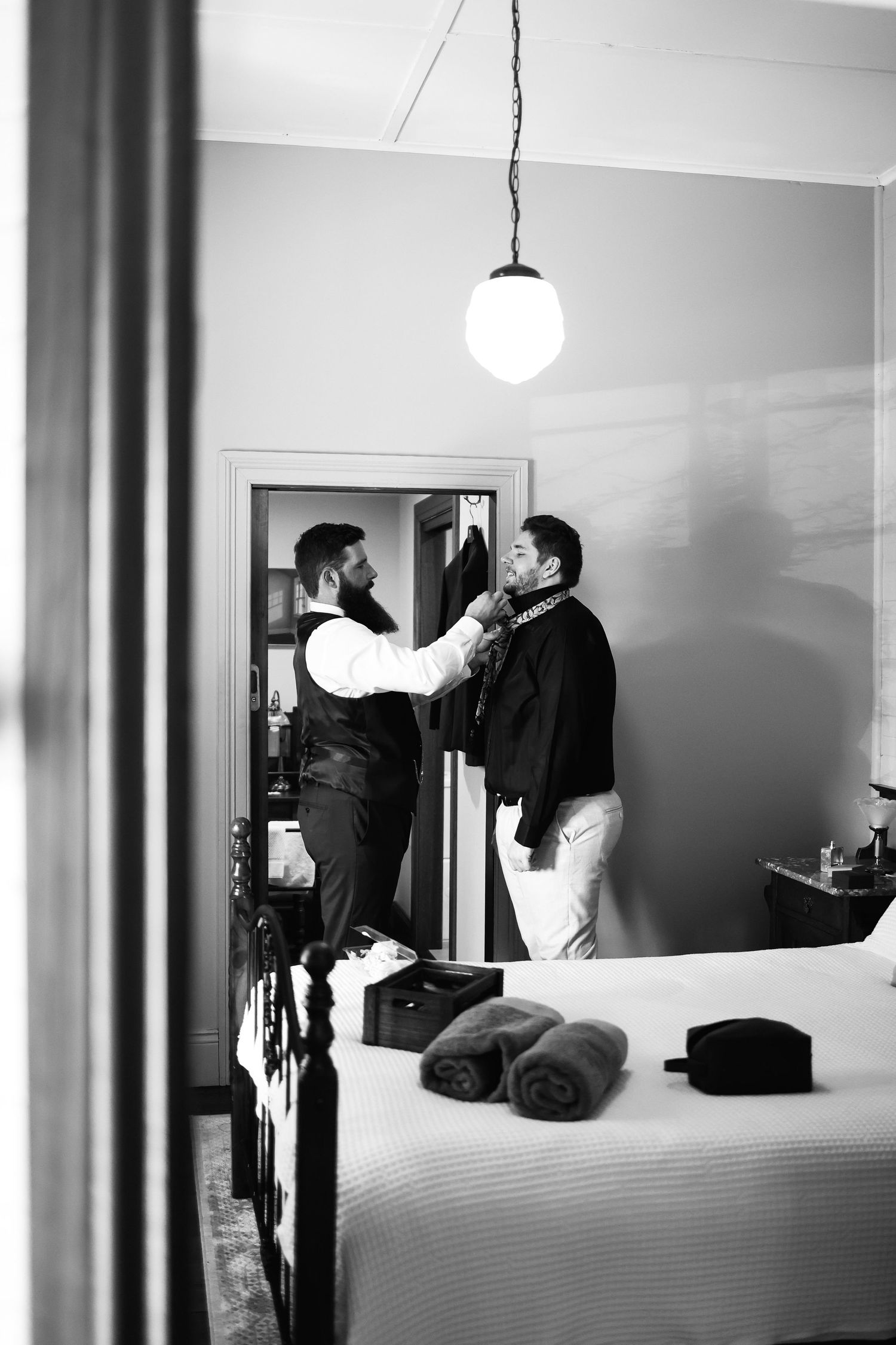 Groomsman helps groom with tie in bedroom mirror reflection during wedding day preparations.