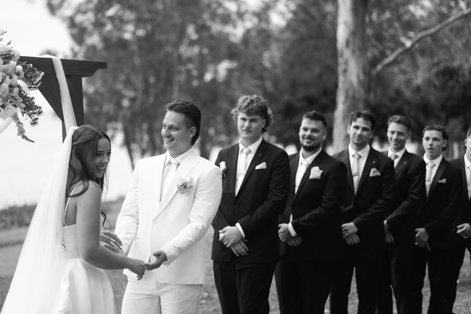 Groom smiling at bride during outdoor wedding ceremony with groomsmen in black suits standing behind.