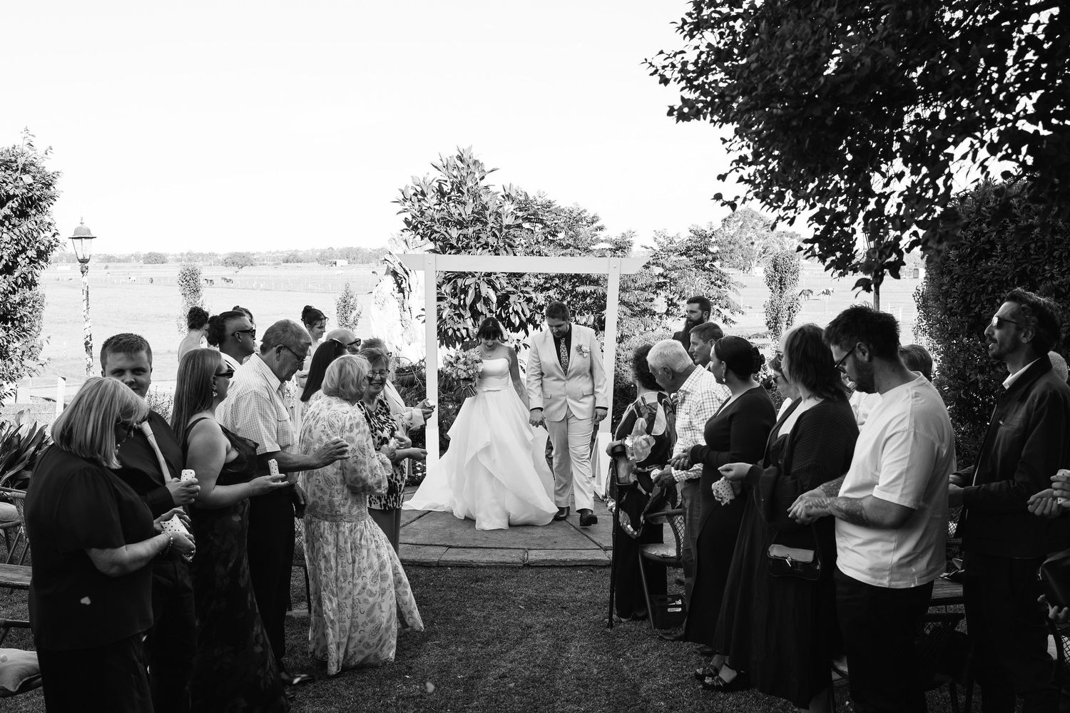 Black and white outdoor wedding ceremony with couple under floral arch surrounded by guests.