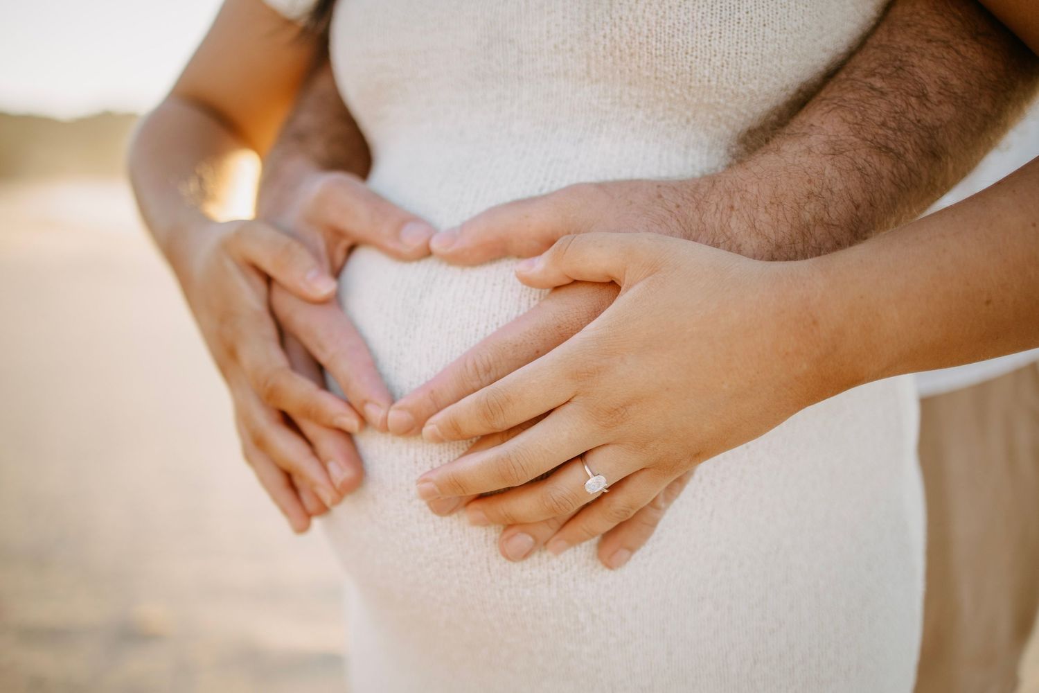 A couple forms a heart shape with their hands on a pregnant woman's baby bump on the beach.