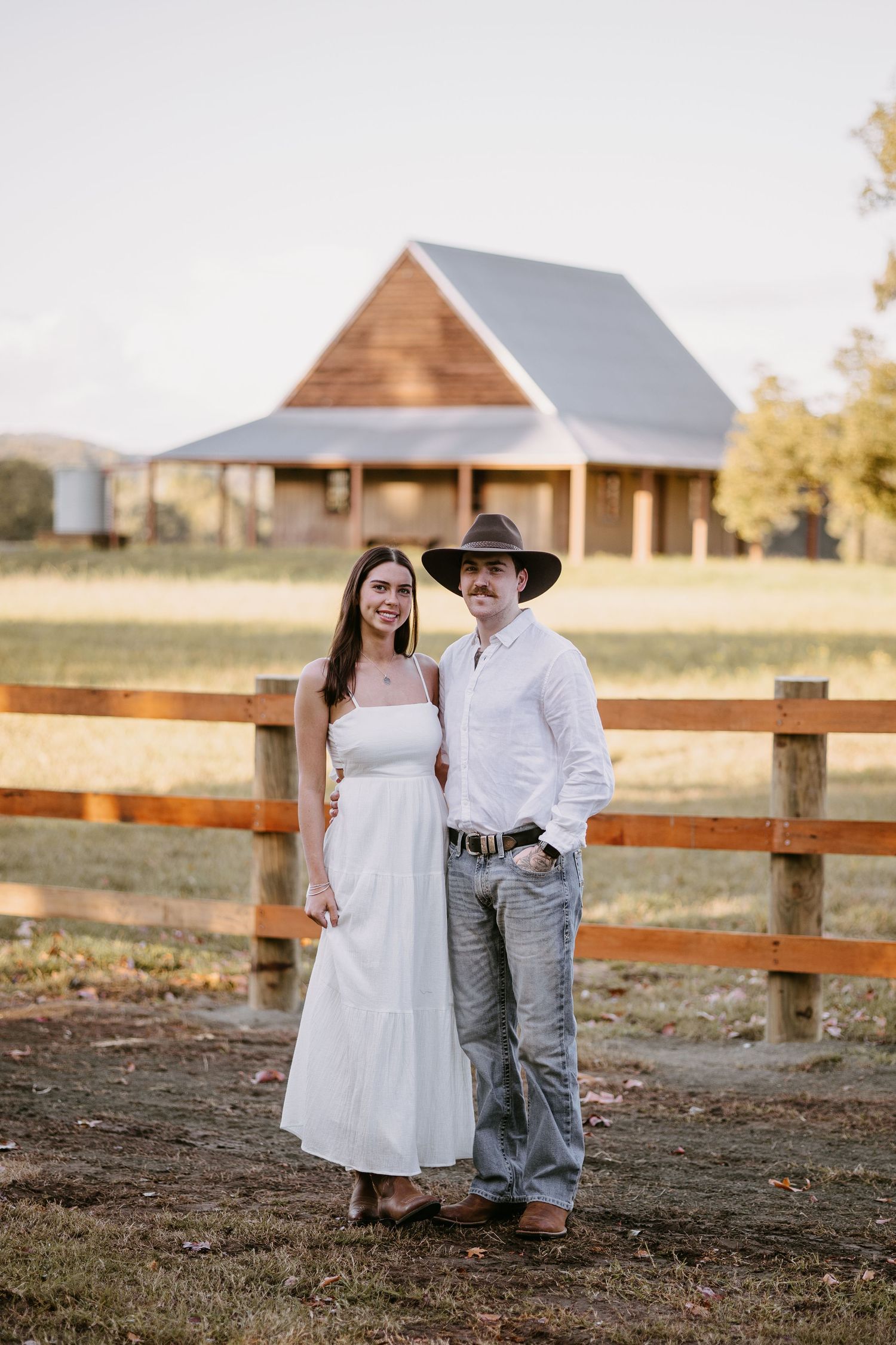 Couple posing together by a wooden fence with a charming rustic barn and green fields behind them.