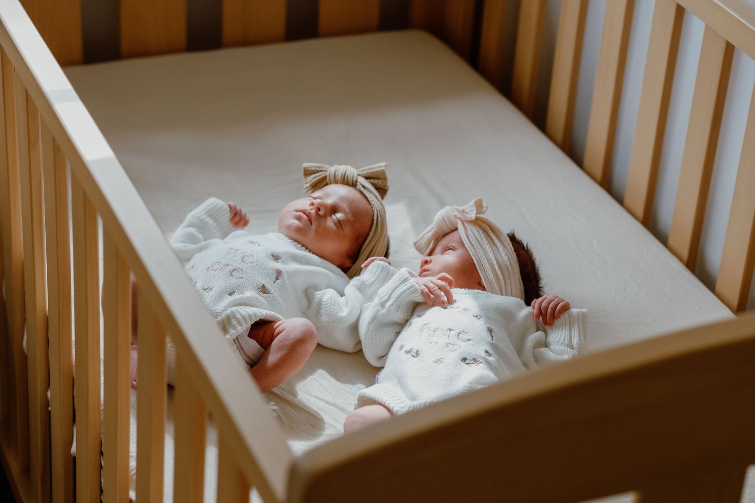 Two sleeping newborn twins dressed in white outfits lying together in a wooden crib.