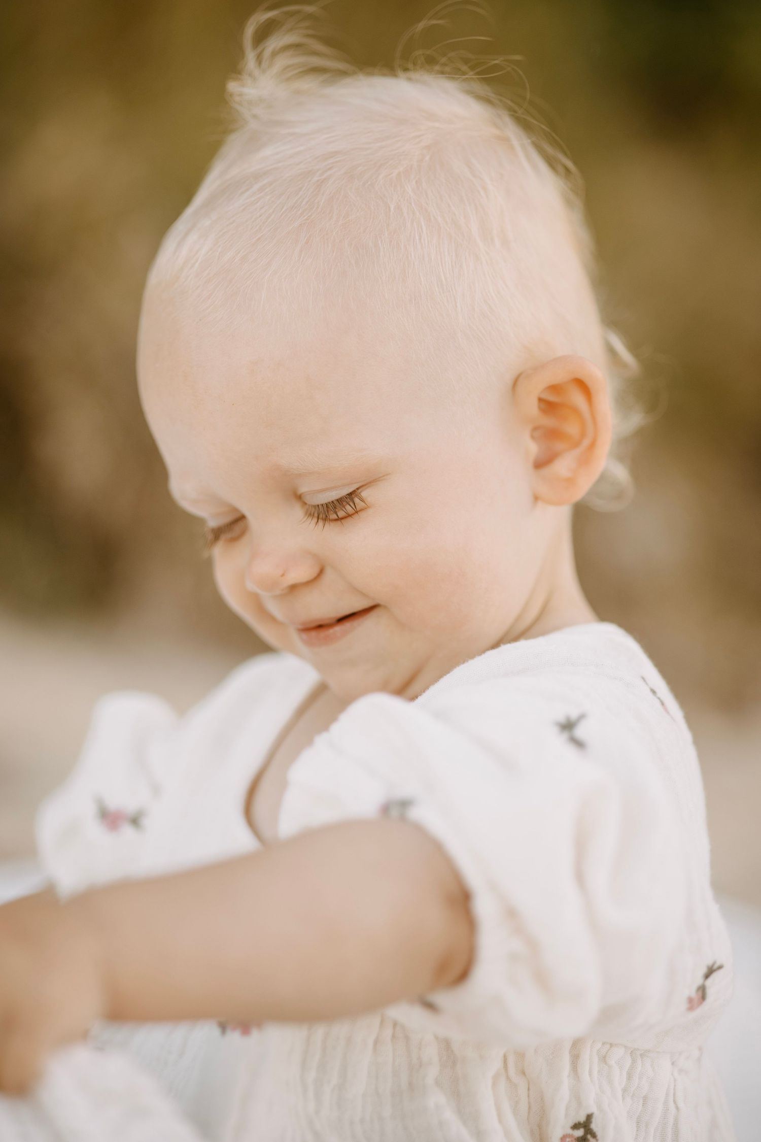 Close-up portrait of a smiling fair-haired baby in white outfit looking downward outdoors.