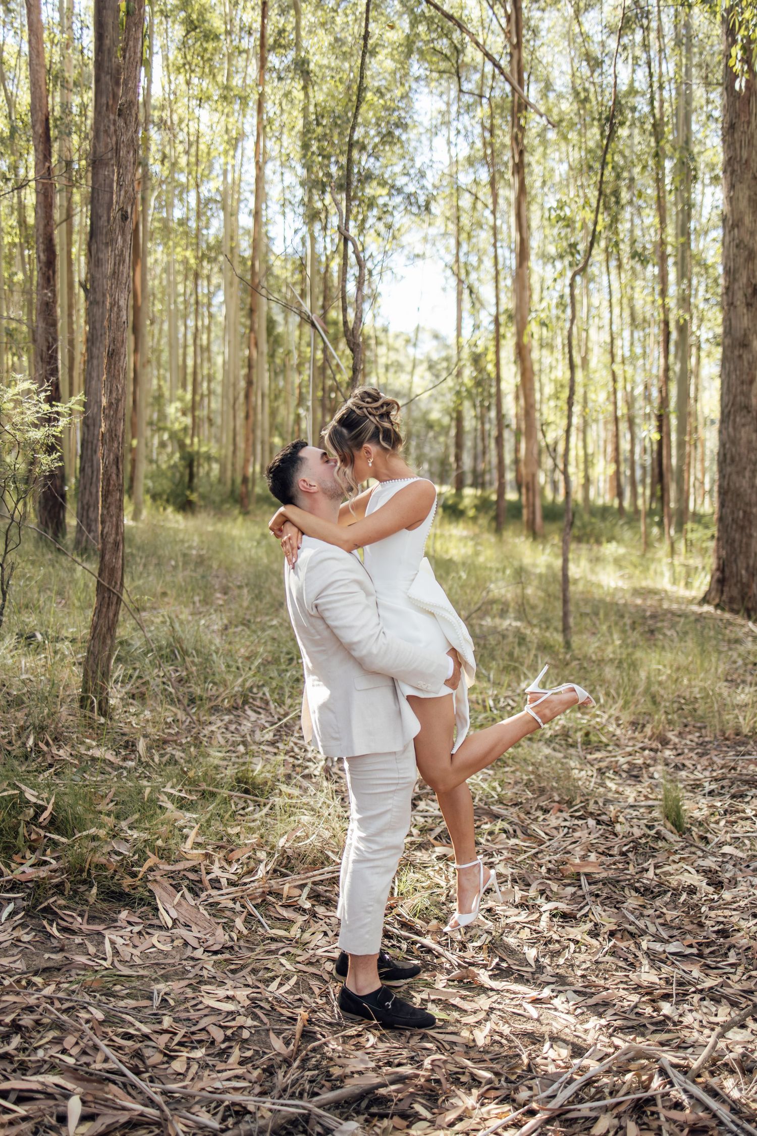 Groom lifting bride in a joyful kiss among tall trees in a forest engagement shoot.
