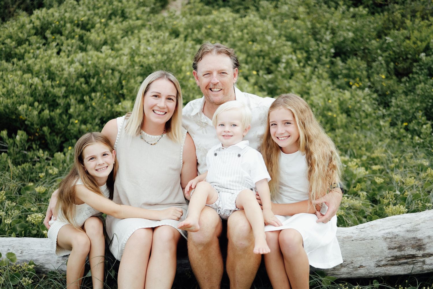 Happy family of five sitting on log with green bushes in background outdoors.
