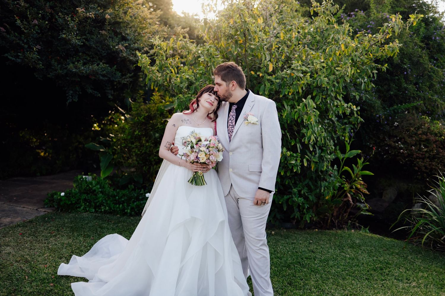Bride and groom share a romantic moment in a lush garden, bride in white gown holding a bouquet, groom in light suit.