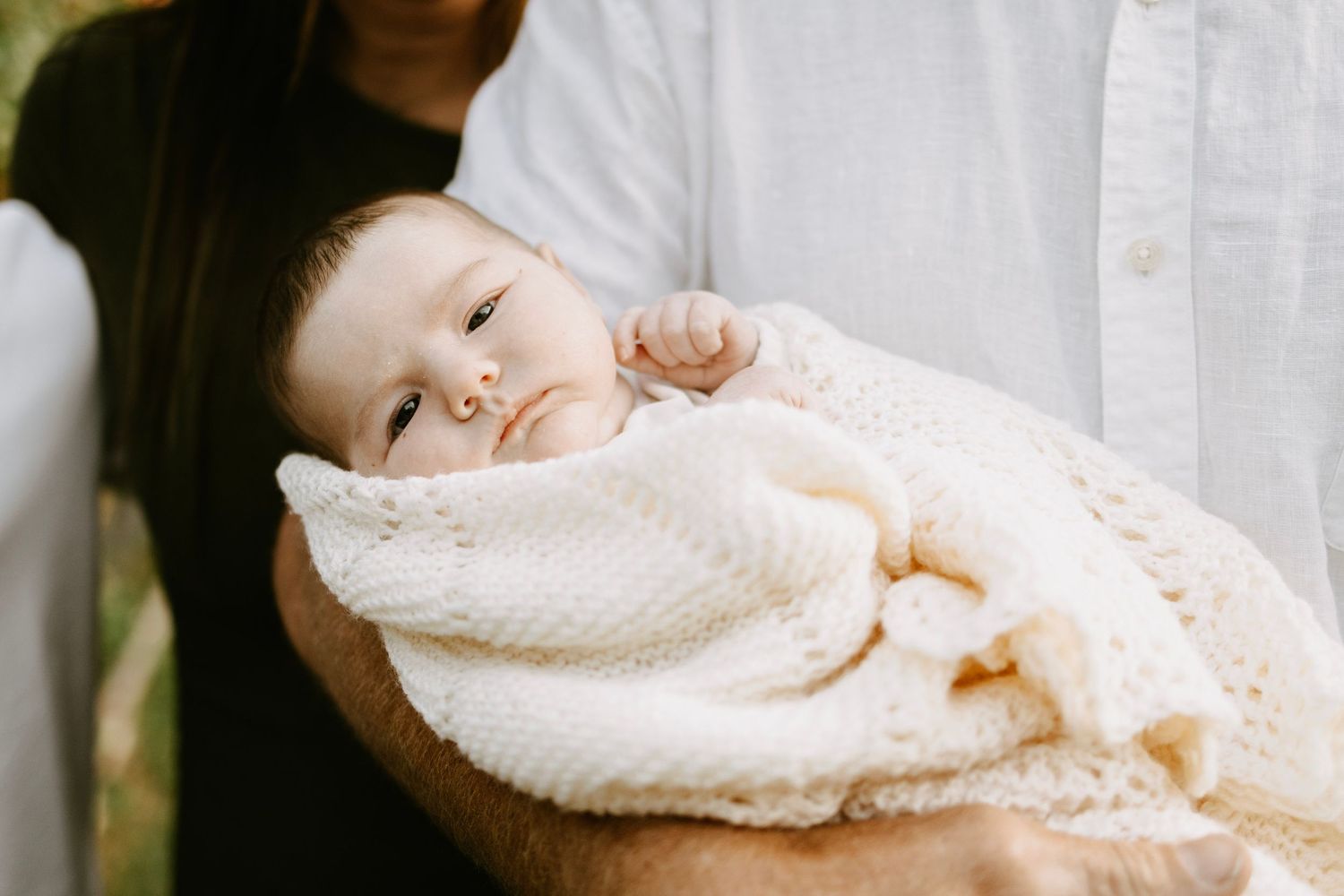 A newborn baby wrapped in a cream knit blanket being cradled in an adult's arms outdoors.