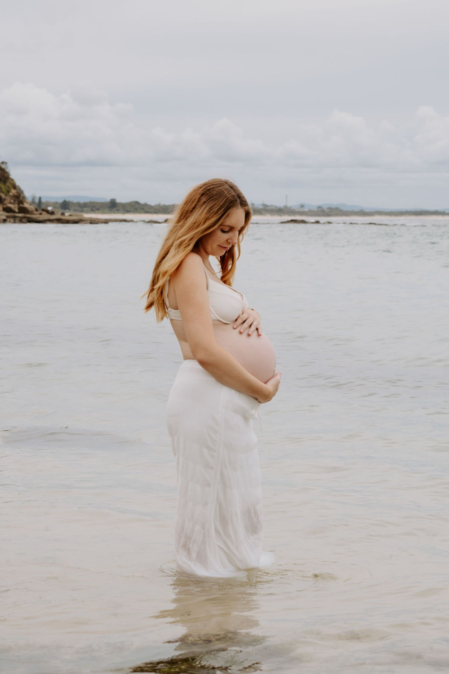 Pregnant woman in white dress standing in shallow ocean water cradling her baby bump.