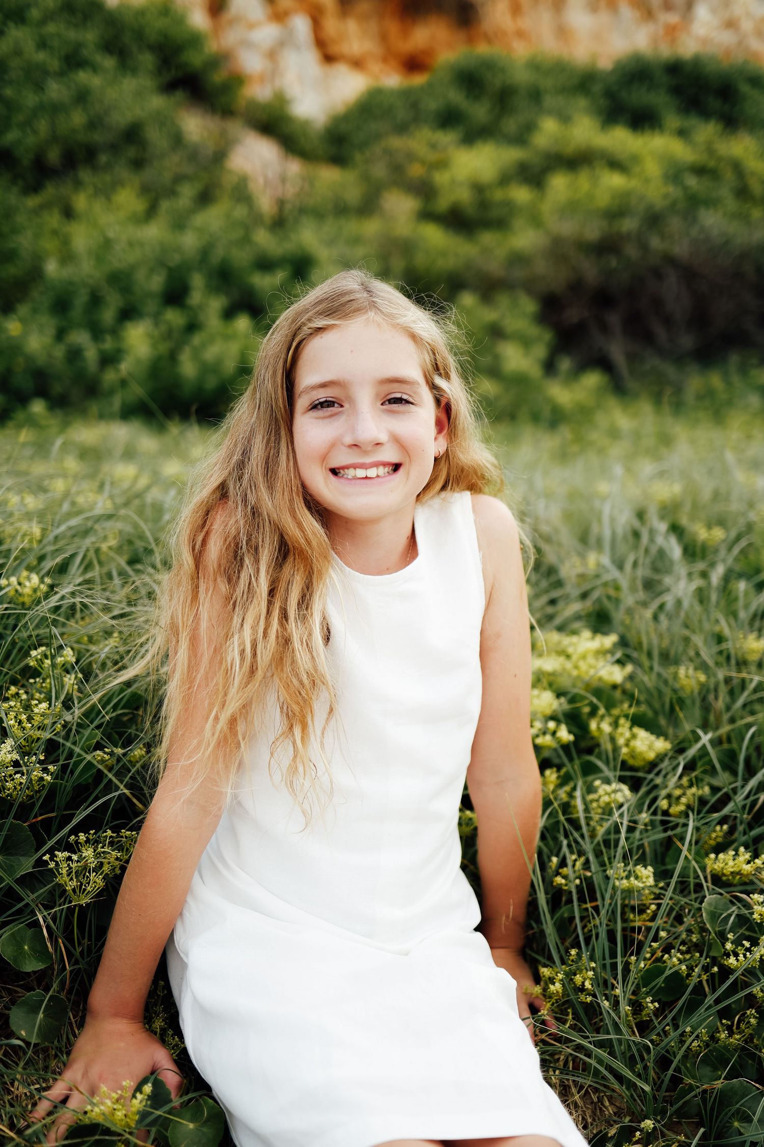 Smiling blonde girl in white dress sitting in green field with yellow wildflowers.