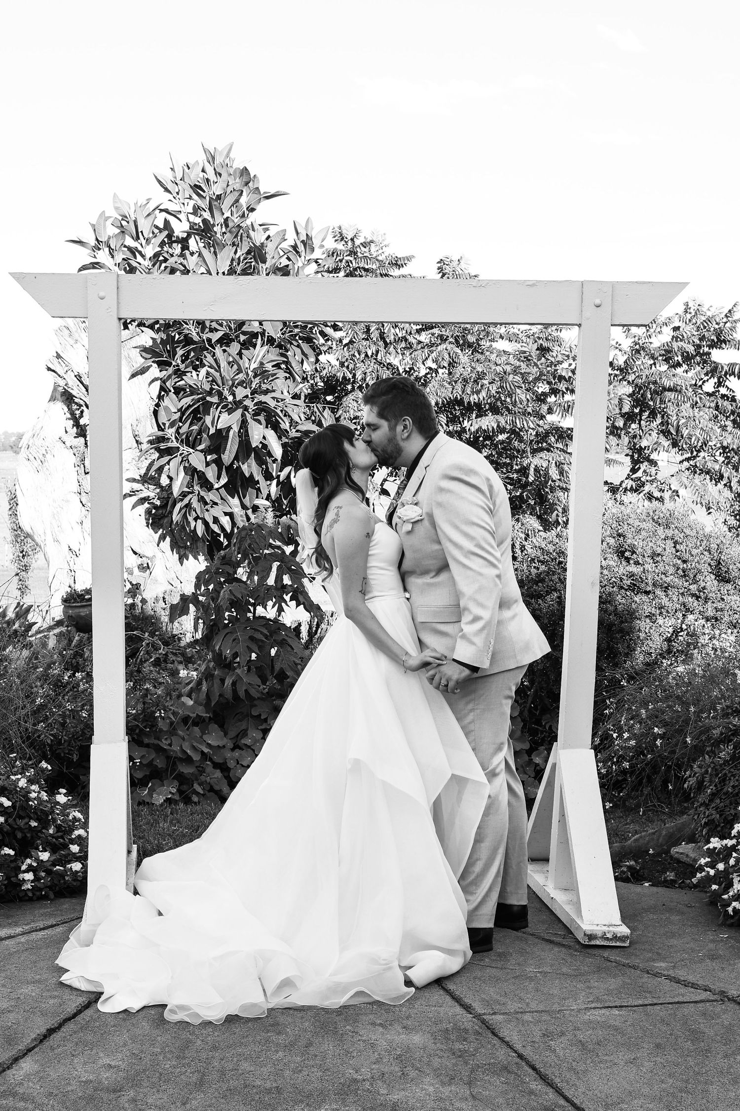 Black and white photo of a bride and groom sharing a kiss under a white wooden arch surrounded by lush greenery.