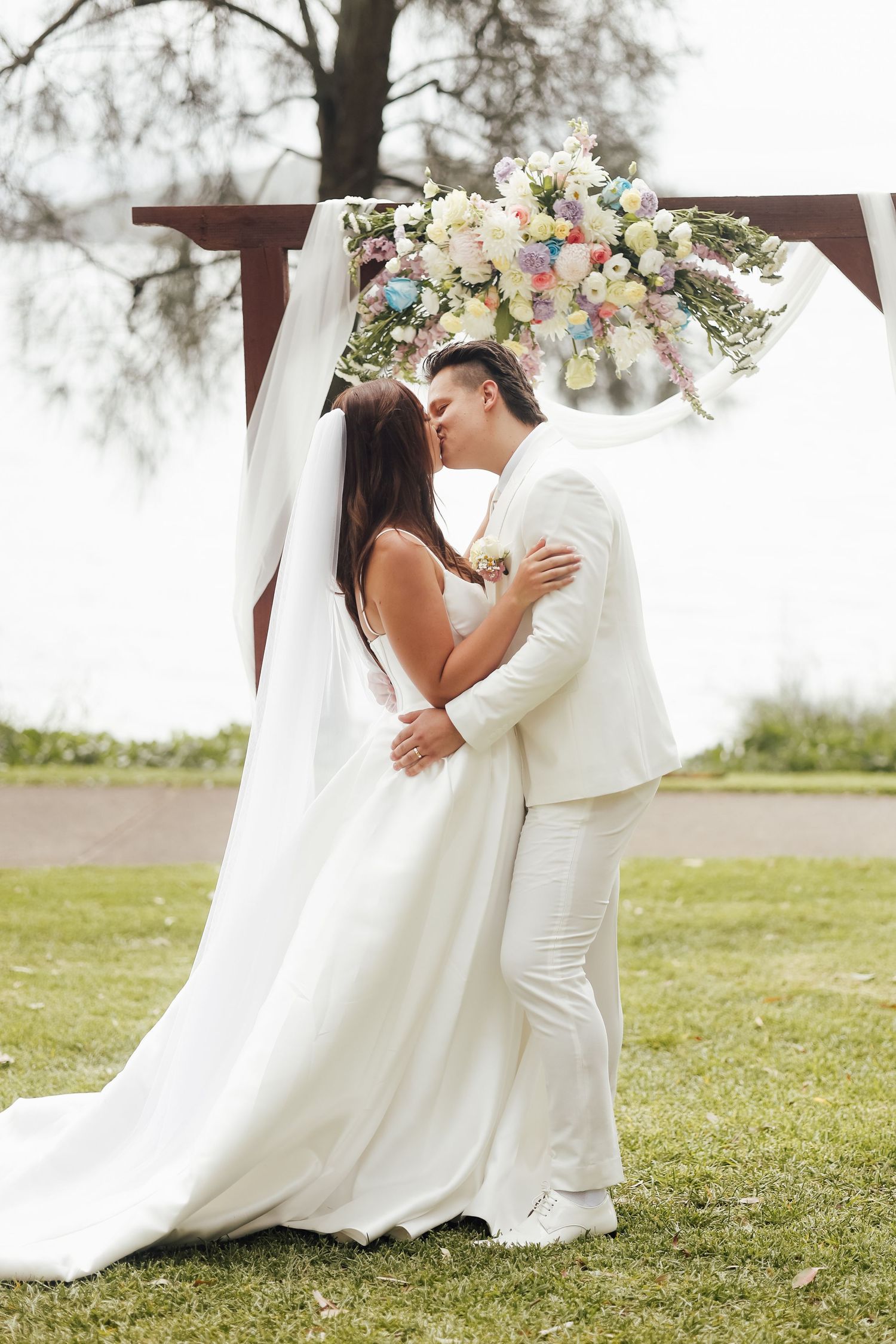 Bride and groom sharing first kiss at outdoor wedding altar decorated with colorful floral arrangement and white draping.