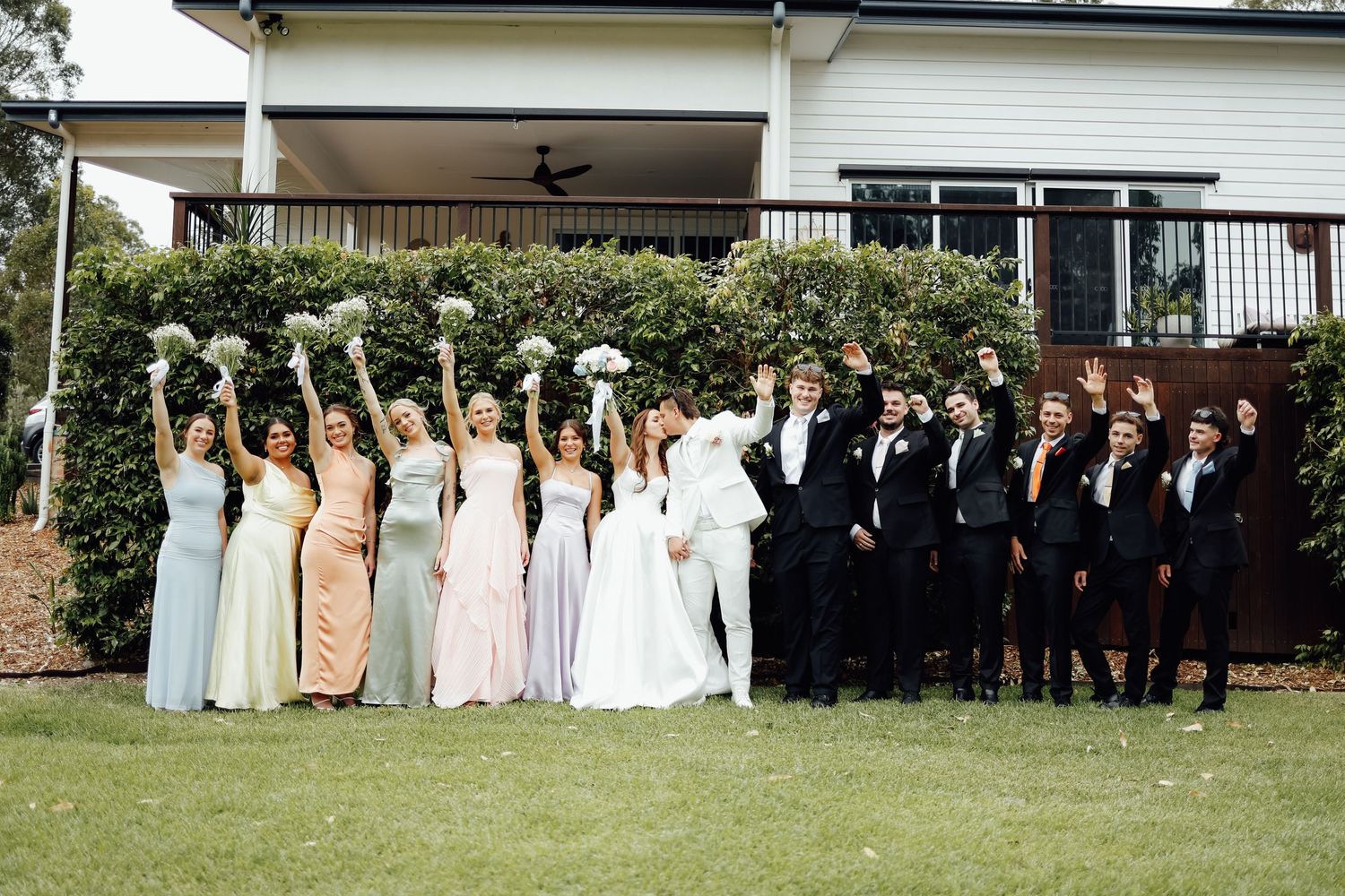 Wedding party poses outside a white house, bridesmaids in pastel gowns holding bouquets, groomsmen in black tuxedos.