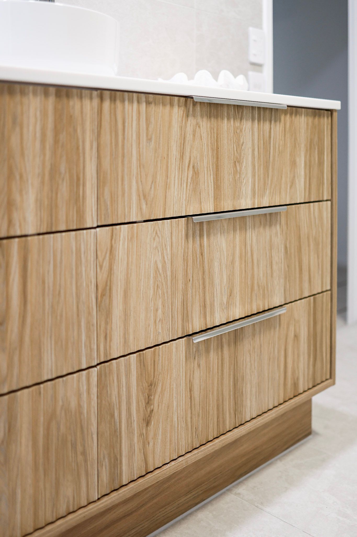 Close-up of a wooden bathroom vanity with three drawers and silver bar handles on a tiled floor.
