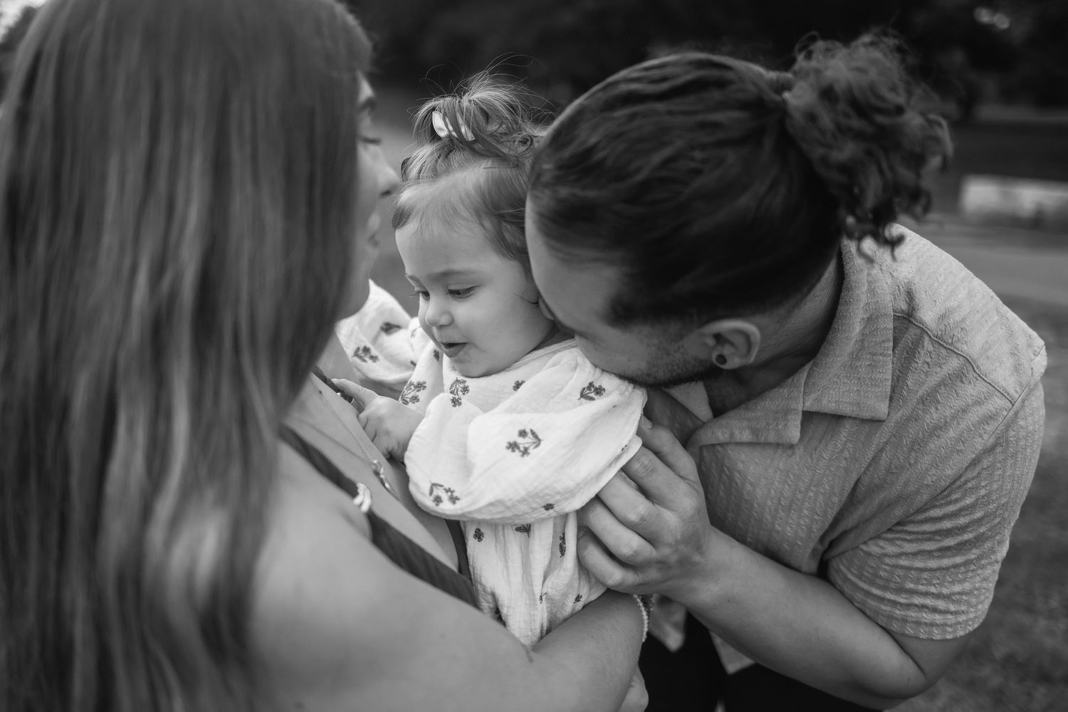 Black and white photo of father tenderly kissing baby girl held by mother outdoors.