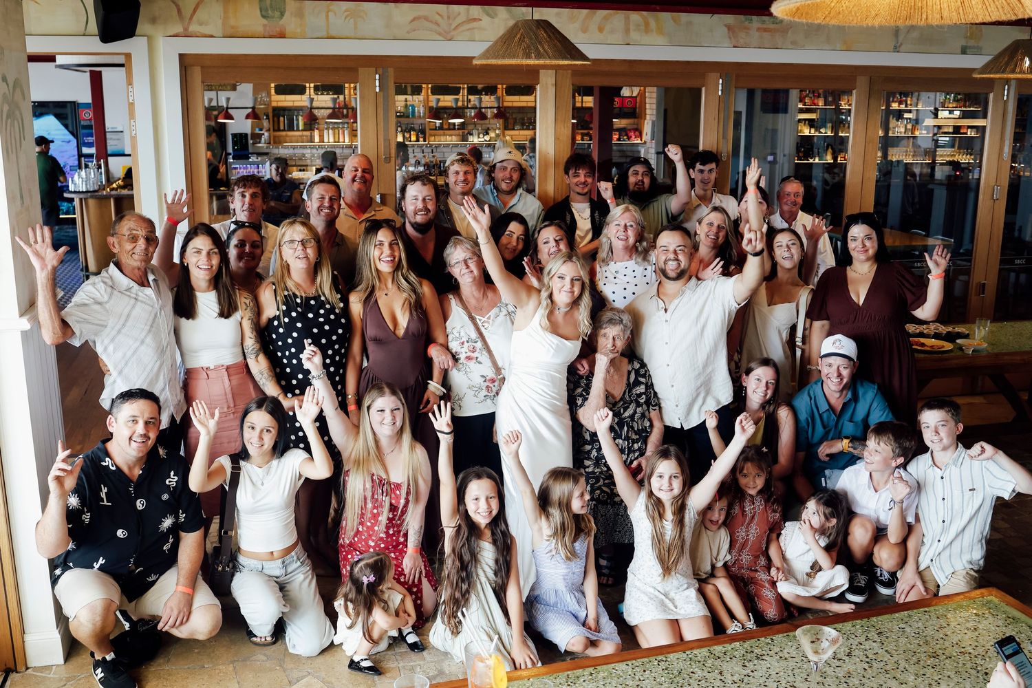 Large wedding group photo with guests in white and neutral tones celebrating indoors.