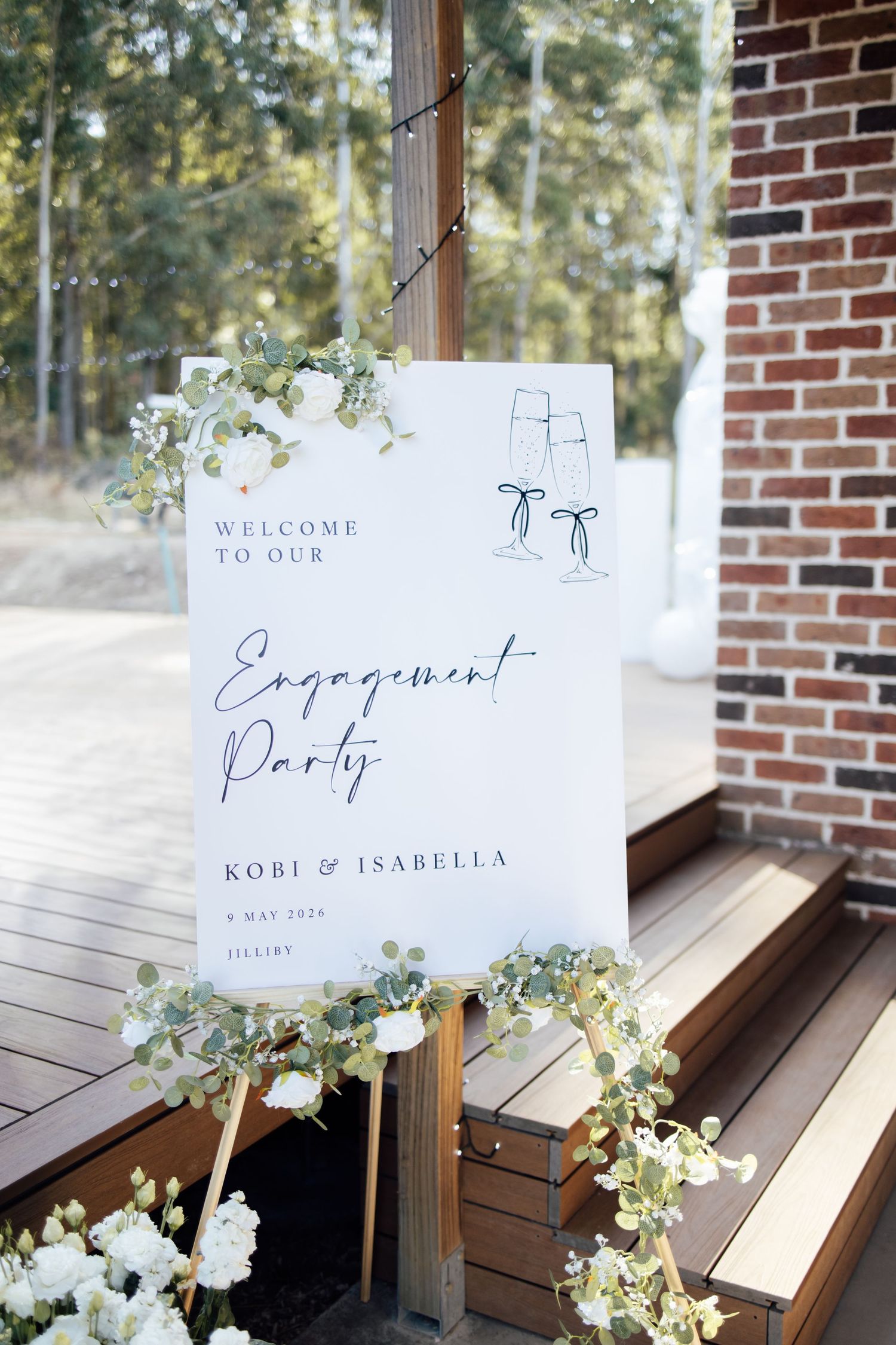 Elegant engagement party welcome sign for Kobi and Isabella, decorated with eucalyptus greenery and white flowers on a wooden deck.