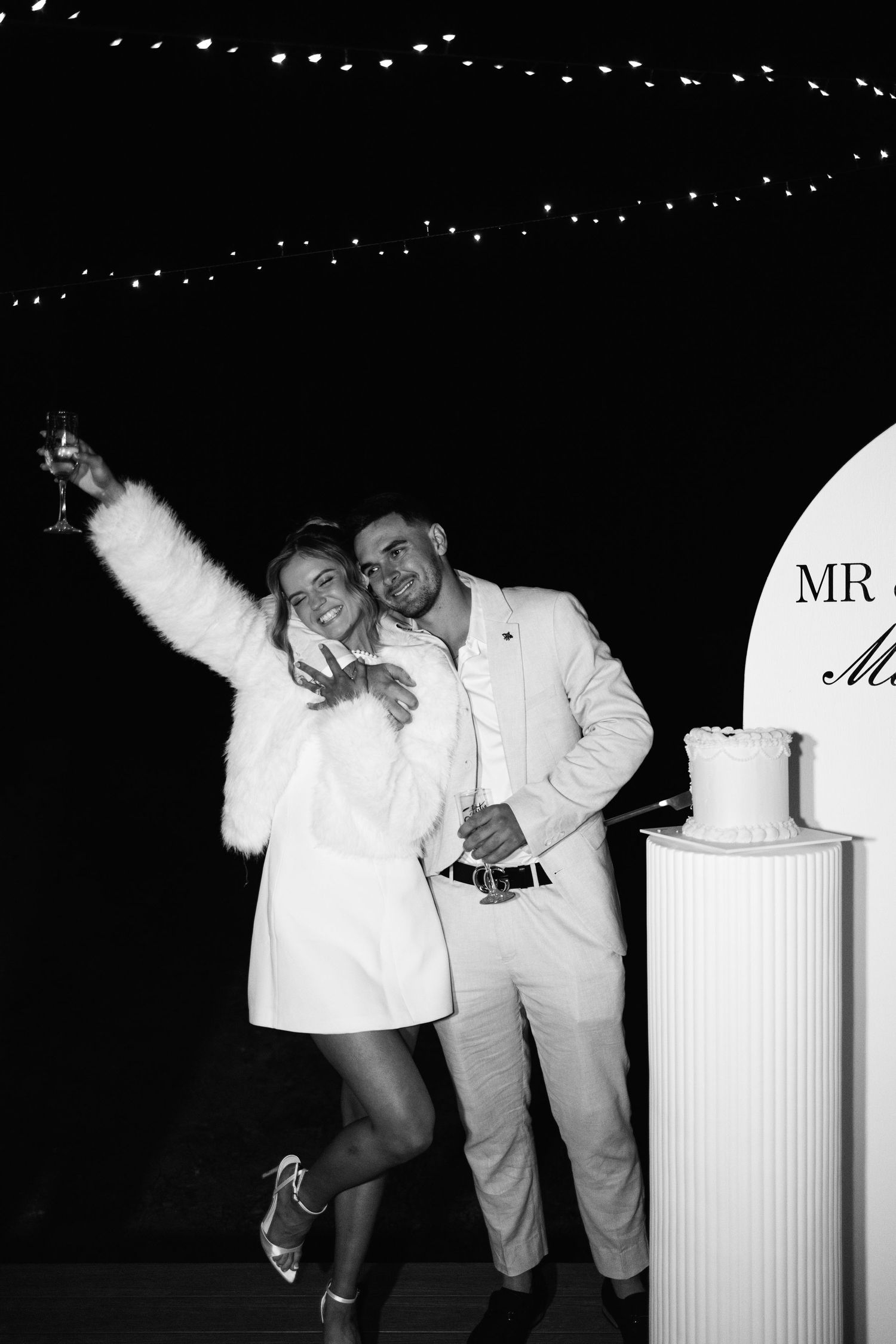 Black and white photo of engaged couple celebrating with champagne beside cake display.