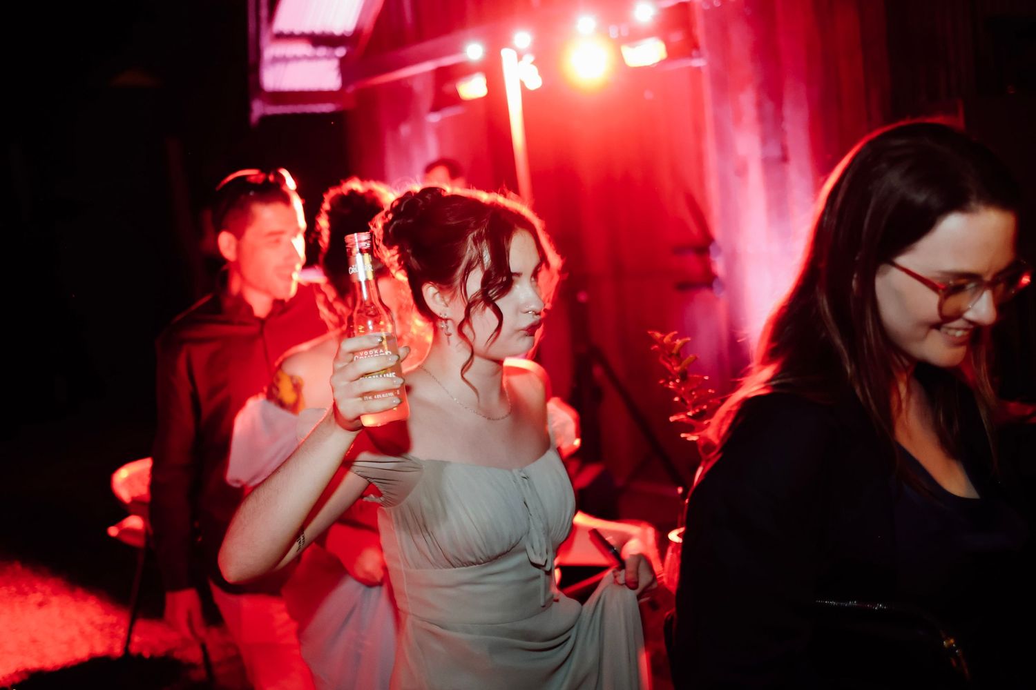 Guests dancing and toasting with beer bottles under red party lights at a festive evening celebration.