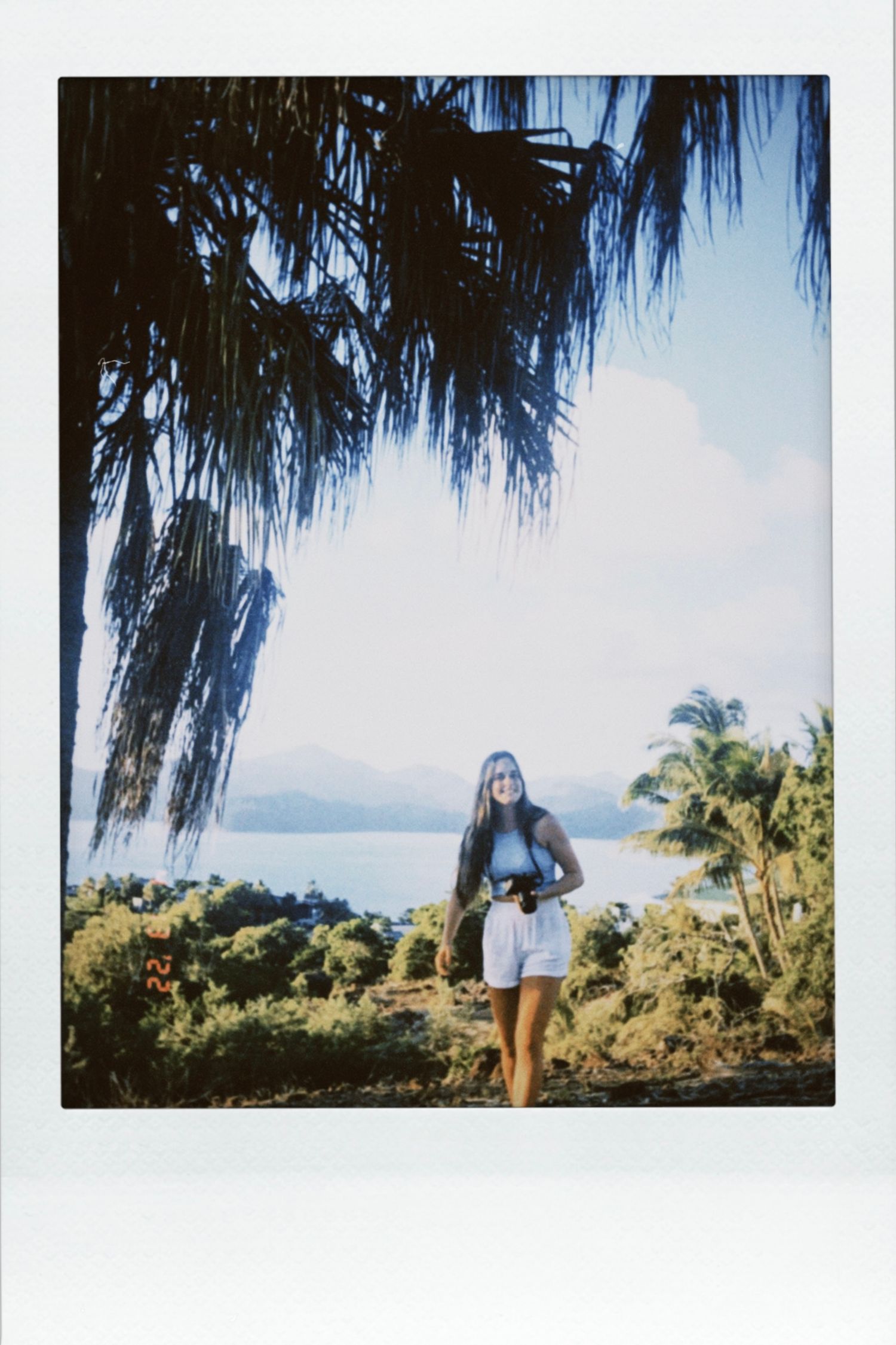 Vintage polaroid of a woman holding a camera under tall palm trees with a tropical lake view.