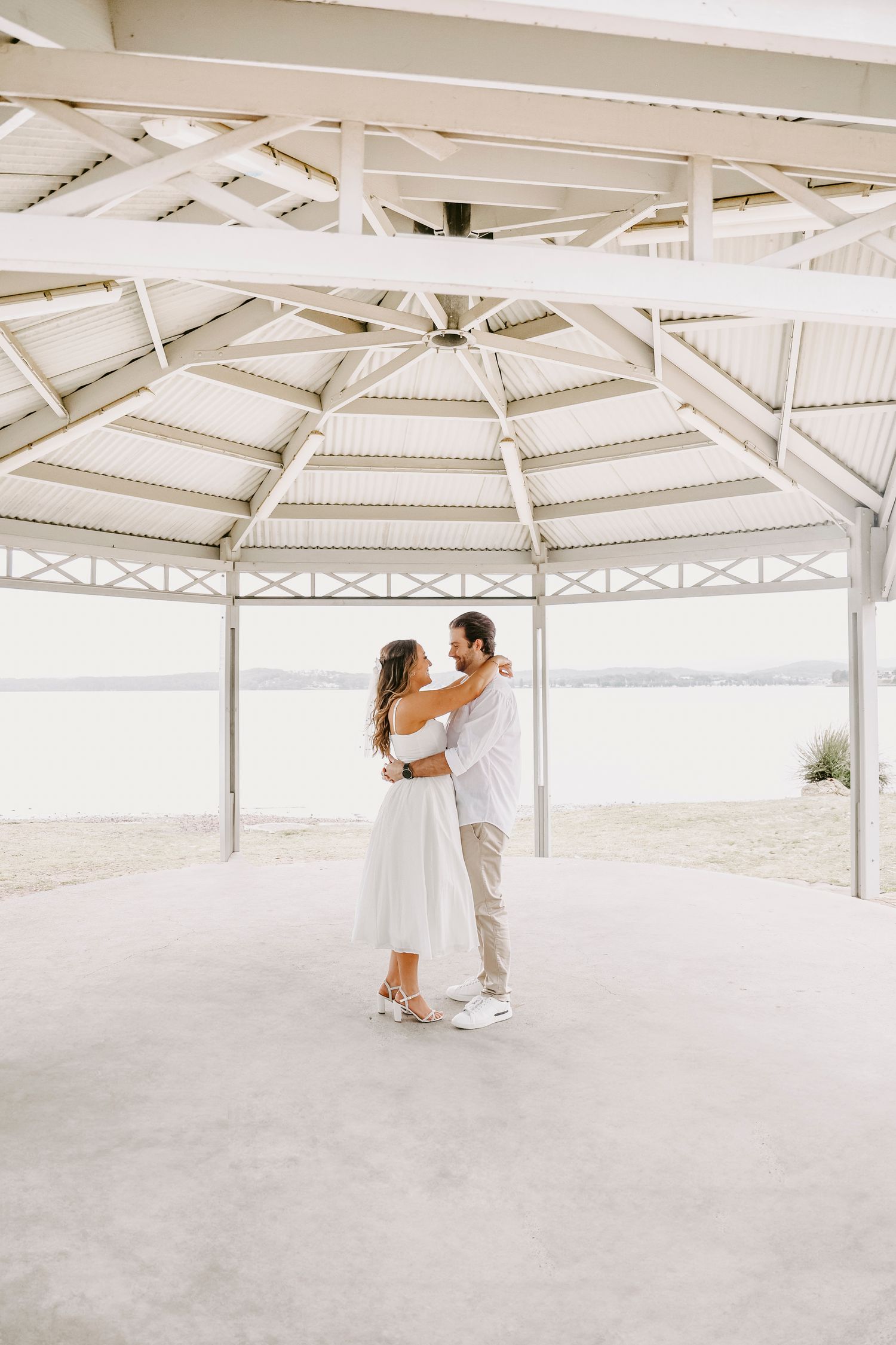Couple dancing and embracing under an open-air white gazebo with ocean views in background.