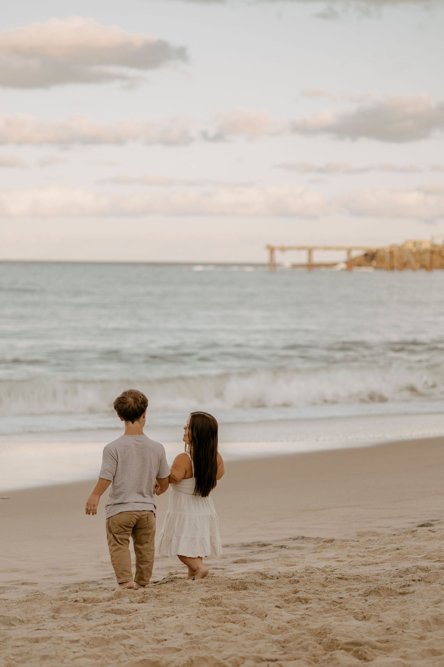 Two children walking hand in hand along a sandy beach toward ocean waves at sunset.