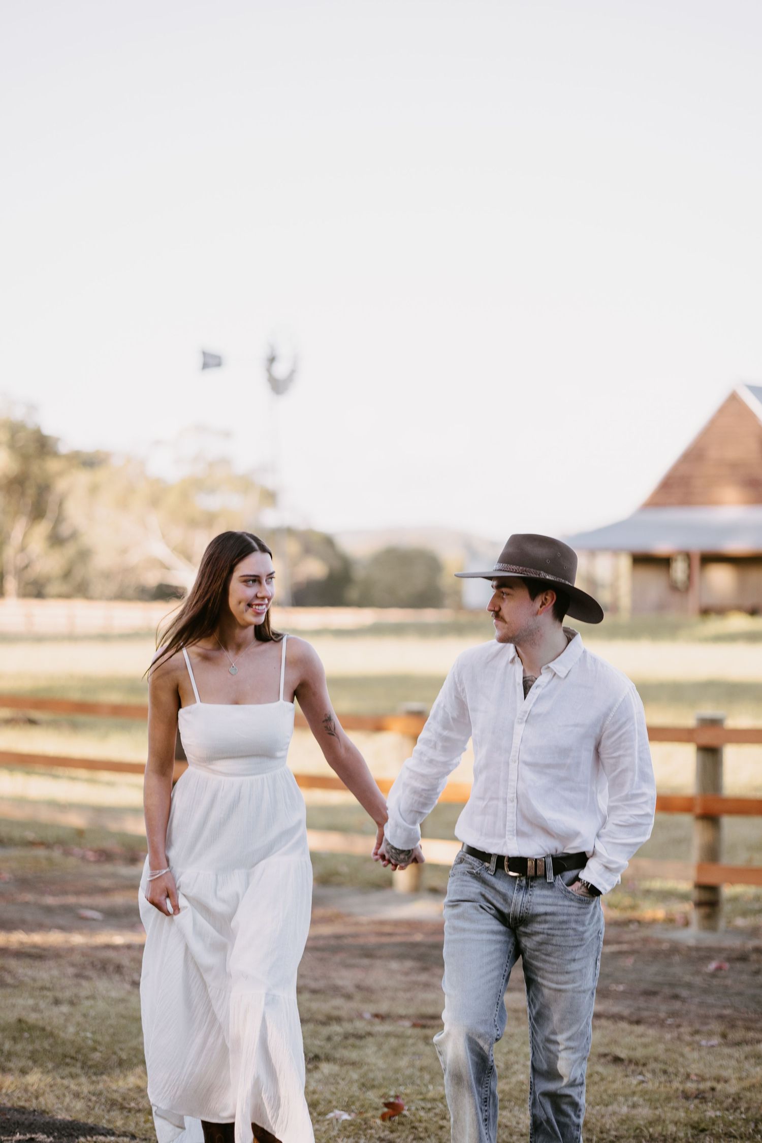 Smiling woman in white dress holds hands with man in cowboy hat walking near rustic farm fence.