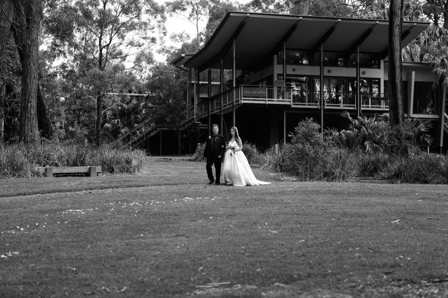 Bride and groom walking across lawn toward elegant venue in black and white photo.