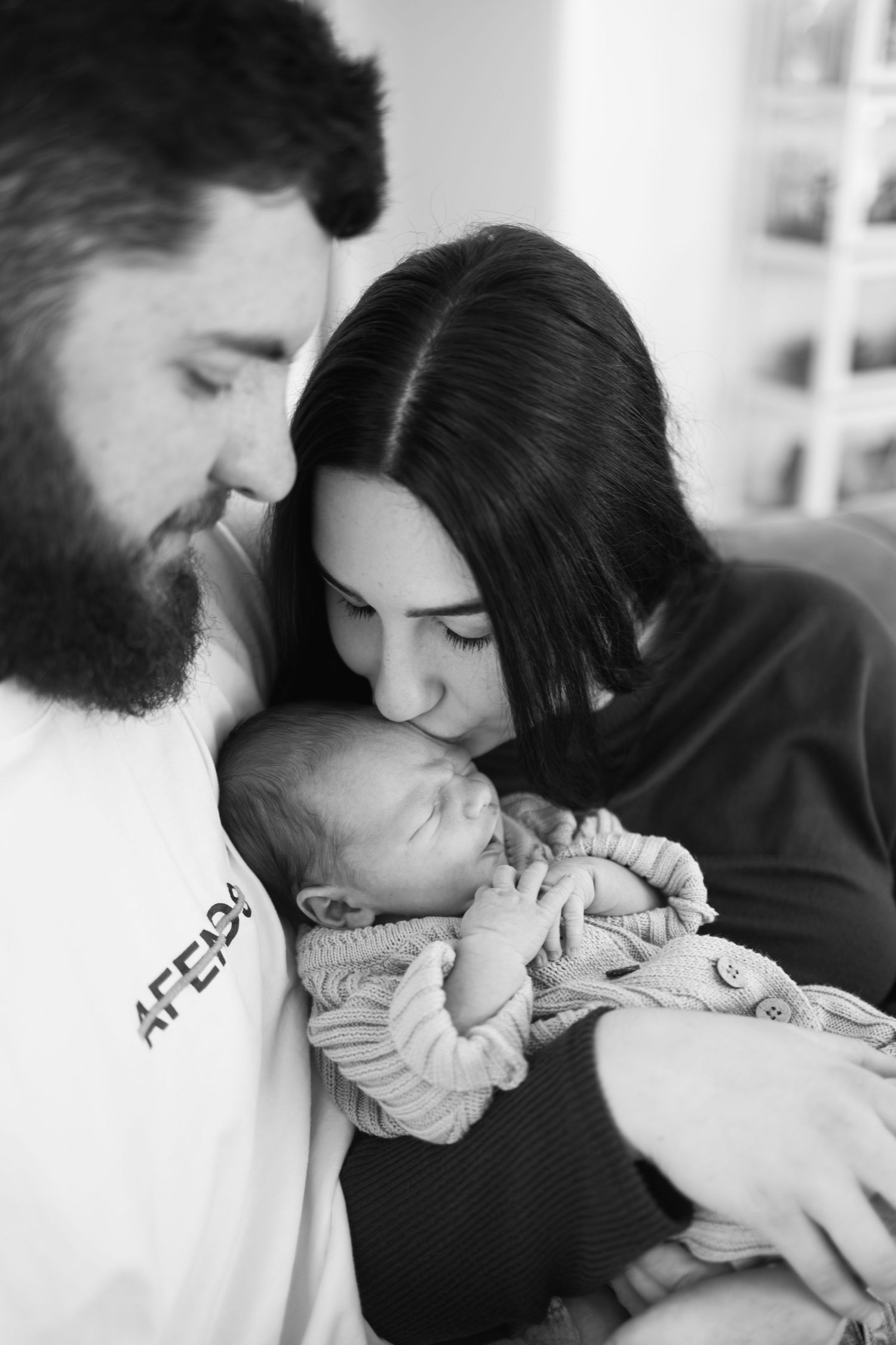 Black and white photo of new parents lovingly holding and kissing their sleeping newborn baby.