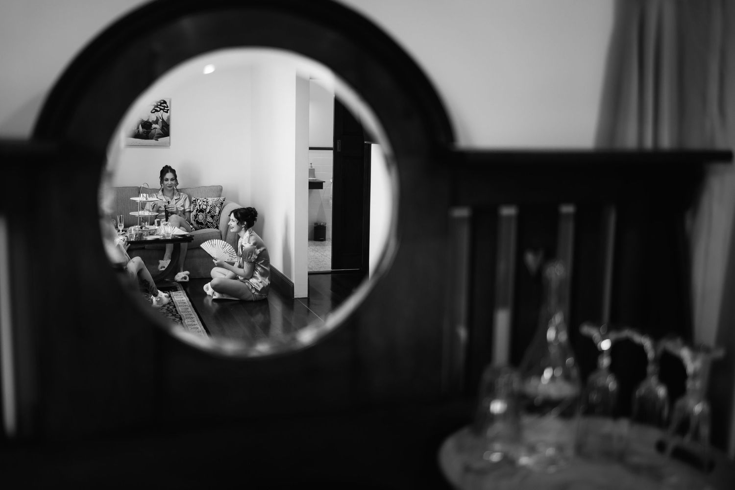Black and white reflection of wedding preparations in an ornate round mirror with glassware in foreground.