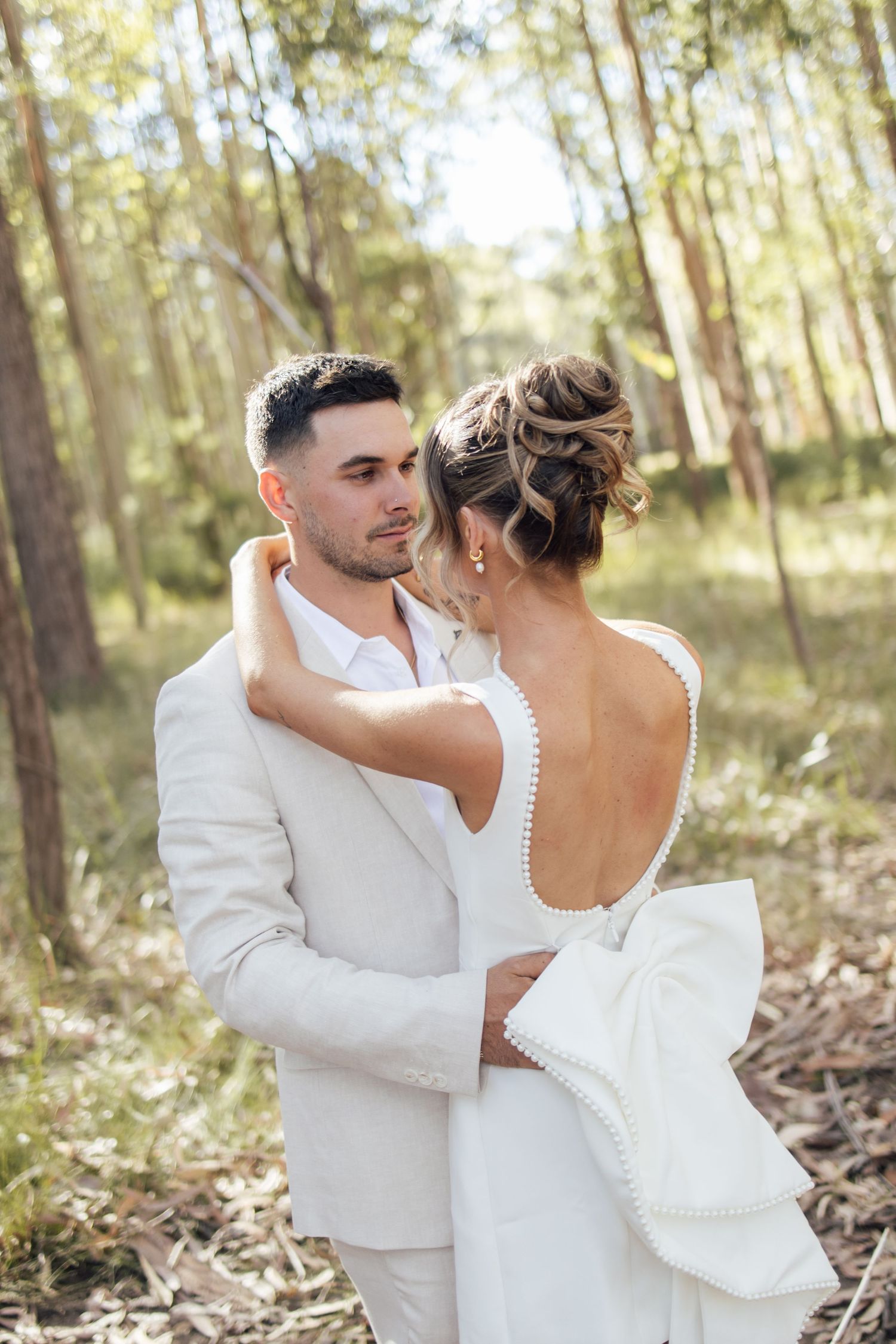 Groom in white suit gazing at bride in backless wedding dress embracing in forest setting.