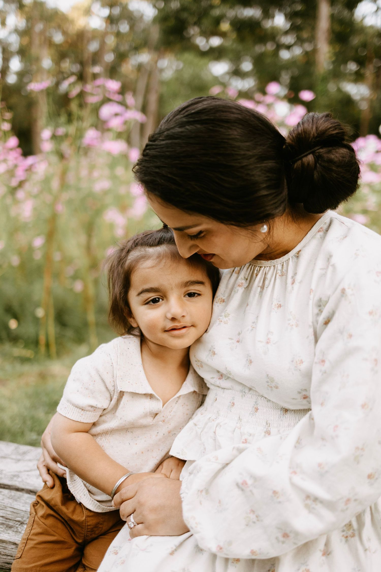 Loving pregnant mother in floral dress embracing young son on bench near pink wildflowers.