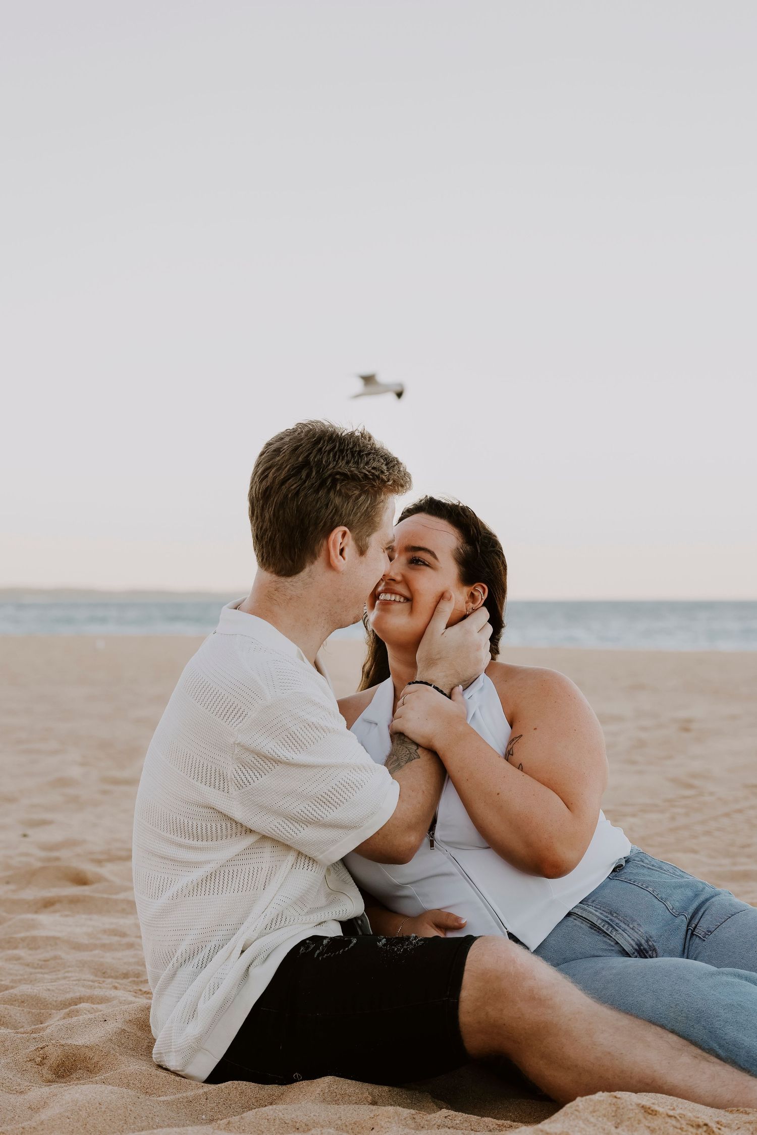 Couple sitting on sandy beach gazing at each other lovingly, man holding womans face.