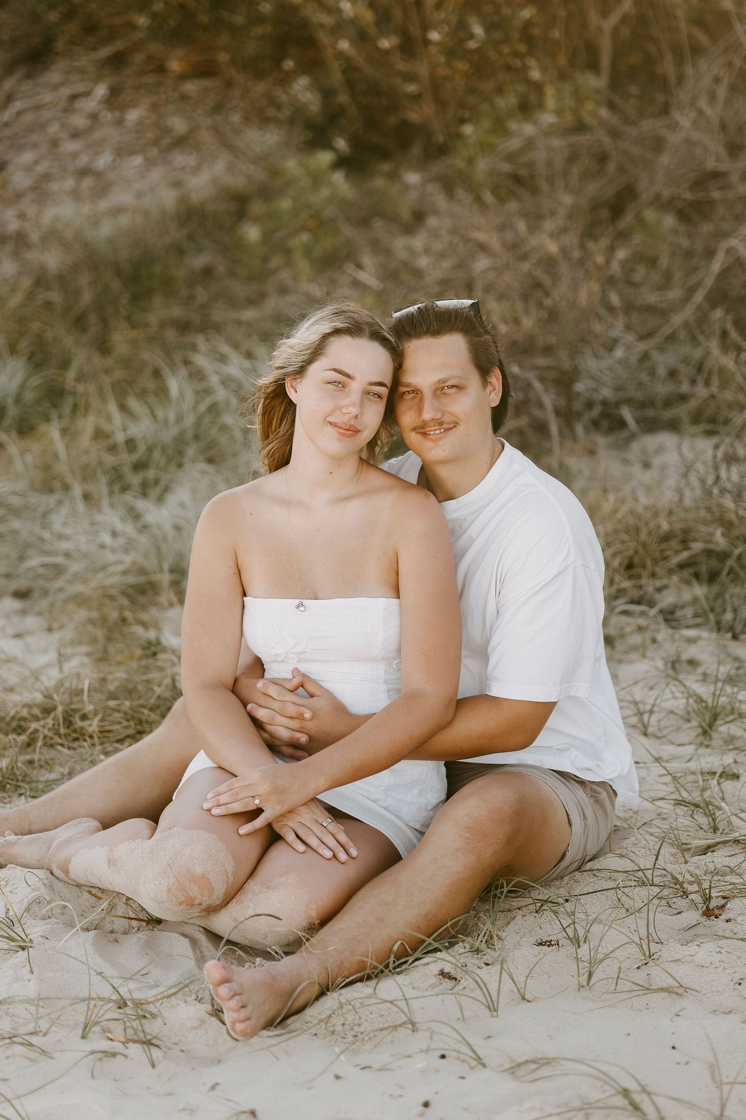 Smiling couple sitting together in sand dunes, woman leaning back into man's embrace at sunset.