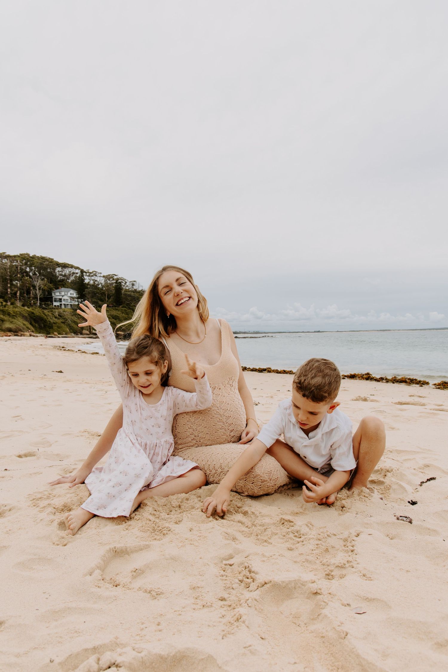 Pregnant woman sitting on sandy beach smiling with two young children on a cloudy day.