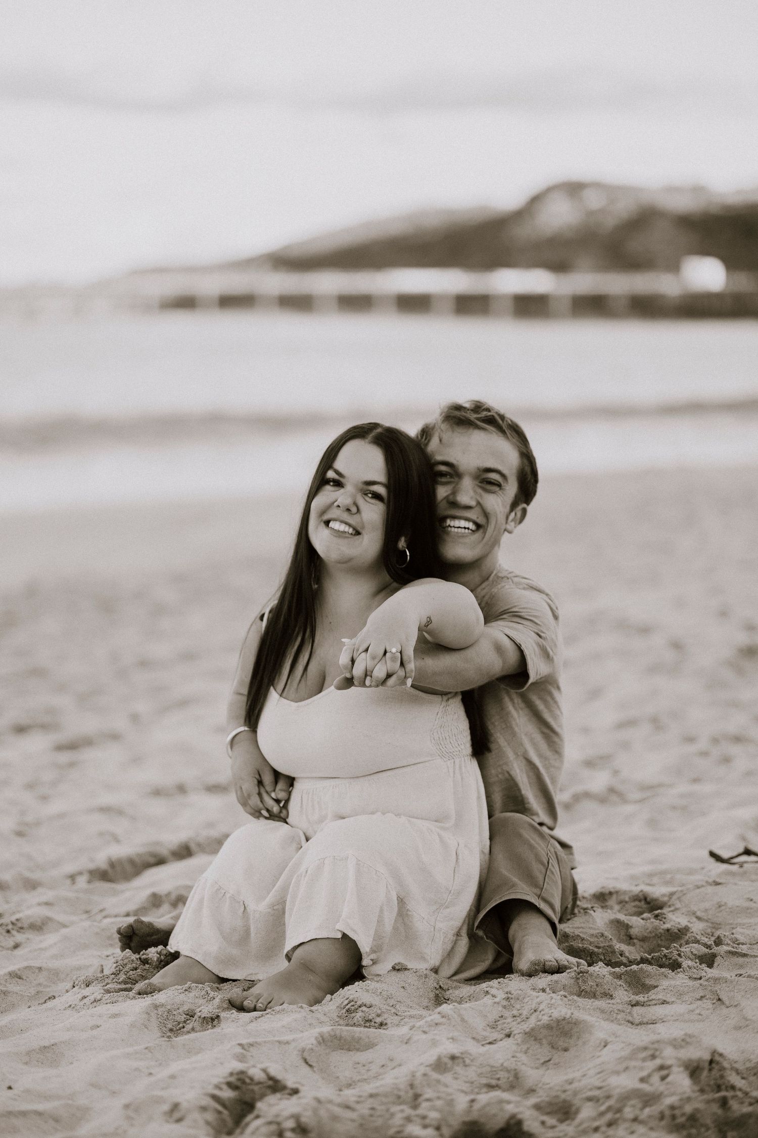 Black and white photo of smiling couple sitting on beach sand, man embracing woman from behind.
