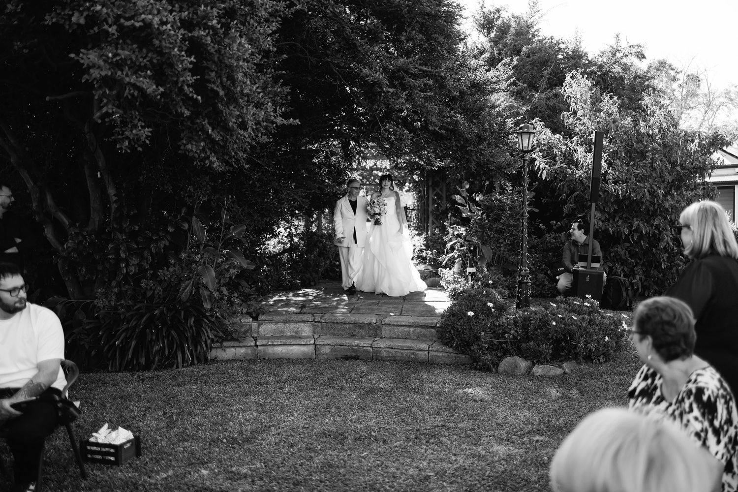 Black and white outdoor wedding ceremony in a garden with guests seated on lawn watching couple at floral altar.