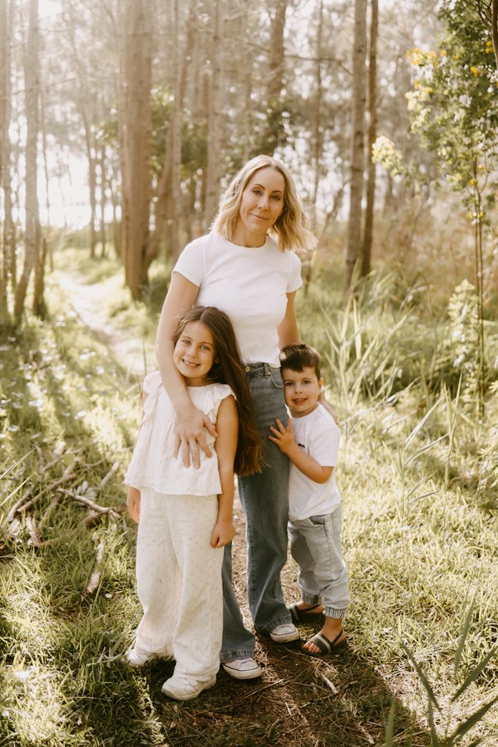 A smiling blonde mother hugs her two young children on a sunlit forest path among tall trees.