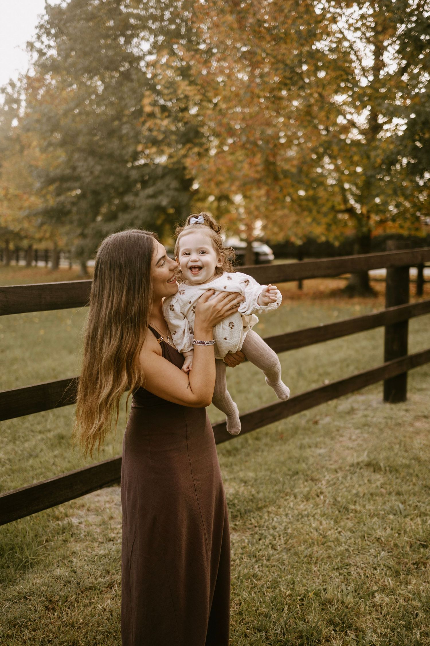 Mother in brown dress kissing smiling baby girl outdoors near wooden fence in autumn setting.