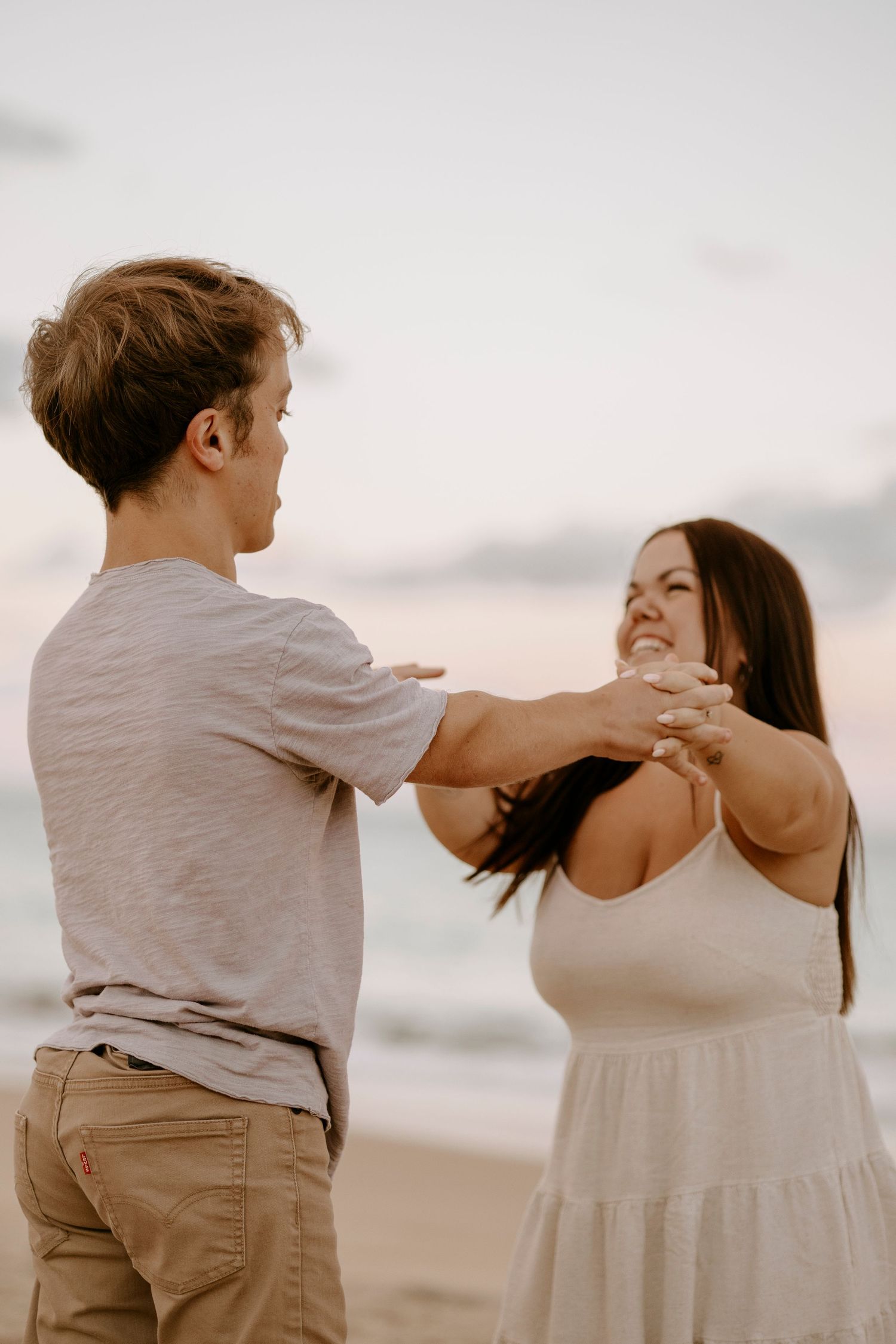 Couple laughing and holding hands on a beach at sunset with ocean waves behind them.